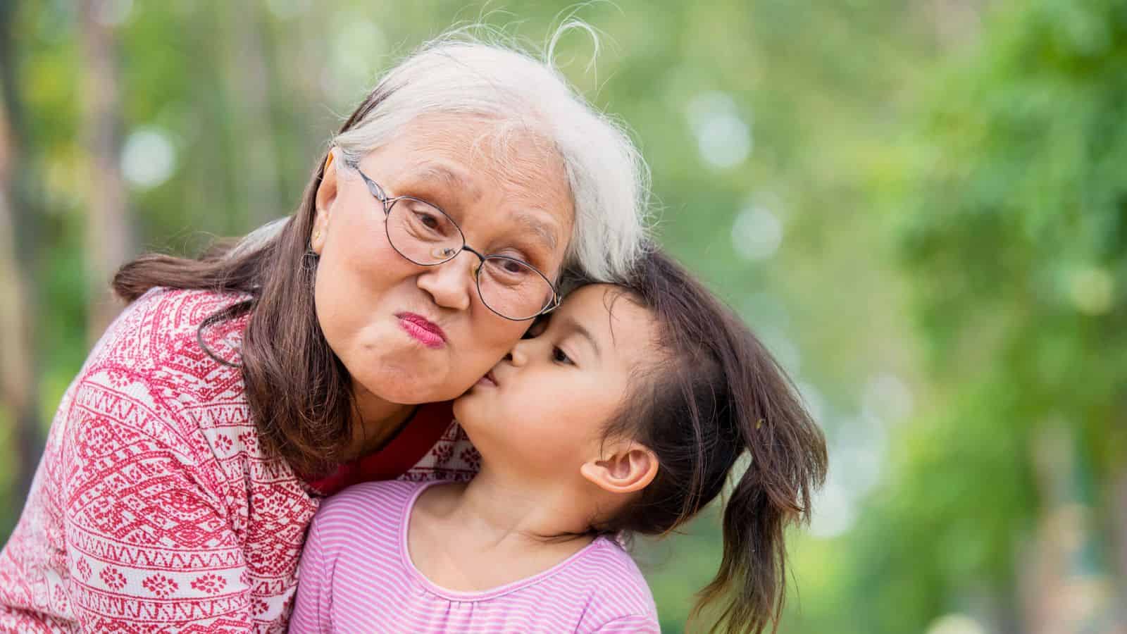 Older woman with glasses being kissed on the cheek by a young girl outdoors, both wearing patterned tops.