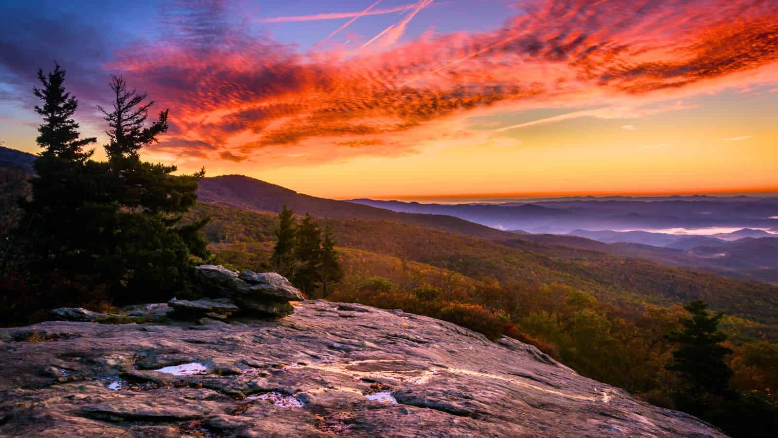 Colorful sunrise over a rocky mountain overlook with trees and rolling hills in the distance.