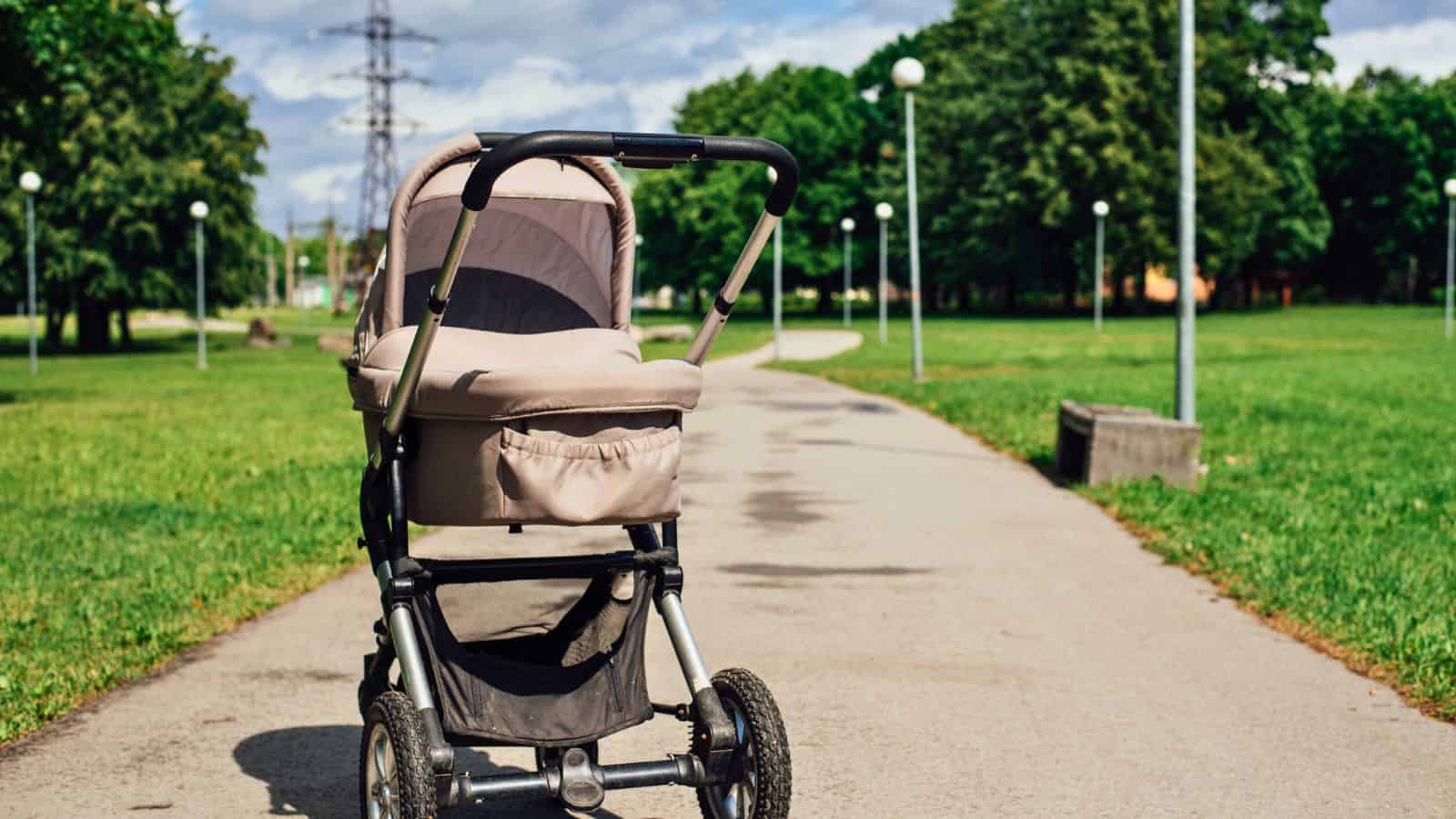 A beige baby stroller stands alone on a paved path in a green park under a partly cloudy sky.