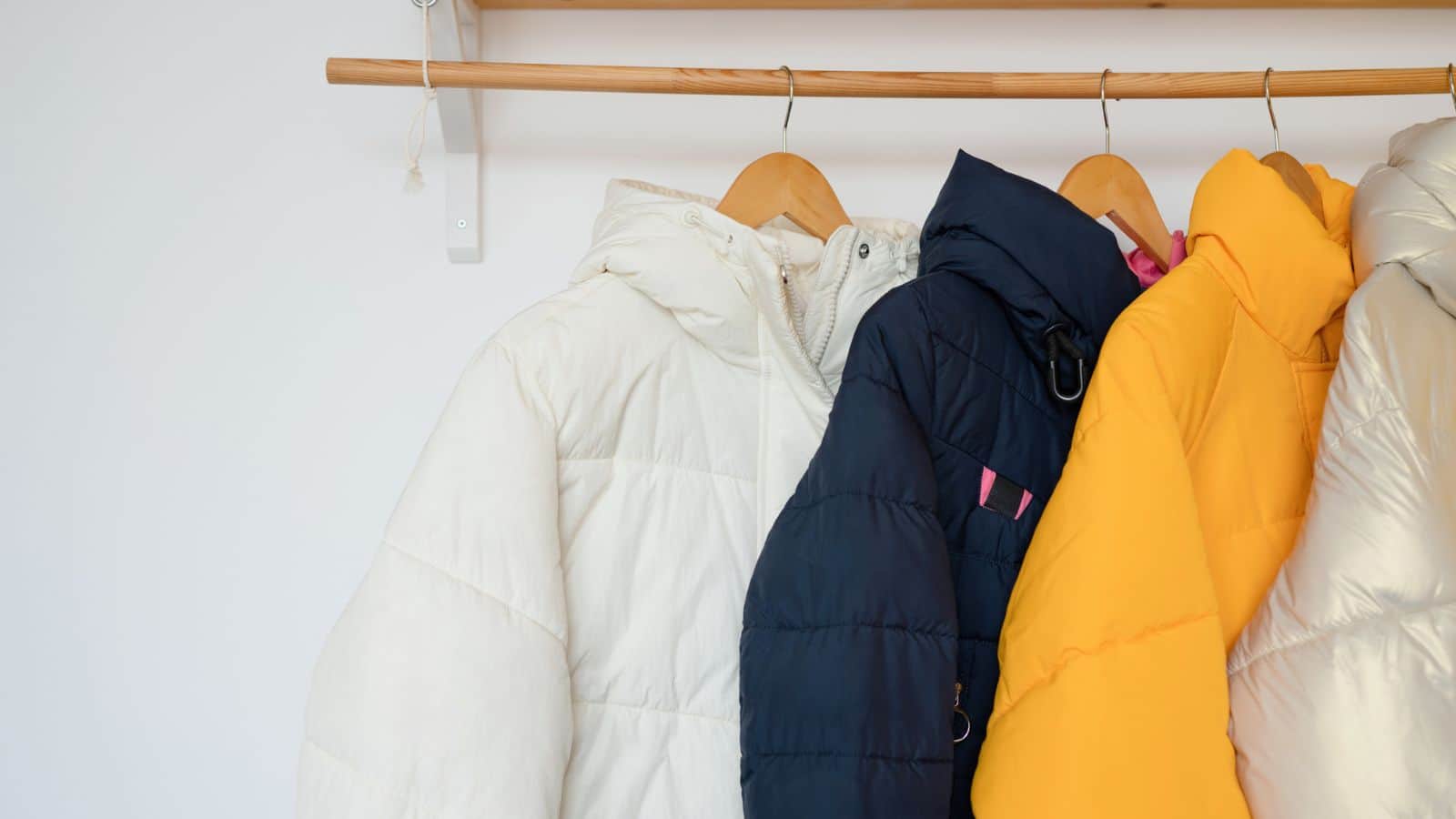 Three puffer jackets—white, navy blue, and yellow—hanging on wooden hangers on a rail.