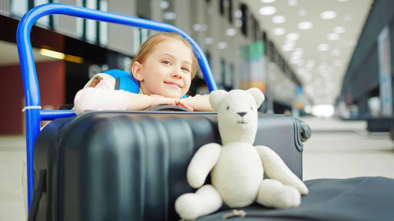 Smiling girl leans on suitcase luggage cart at an airport with a white stuffed bear in the foreground.