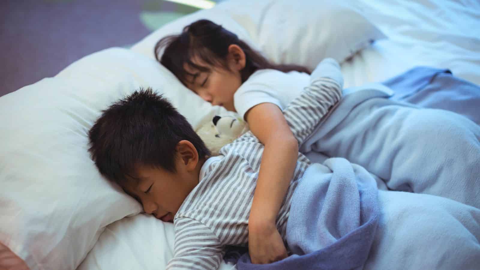 Two children sleeping together in bed, hugging a stuffed animal, covered with blankets.