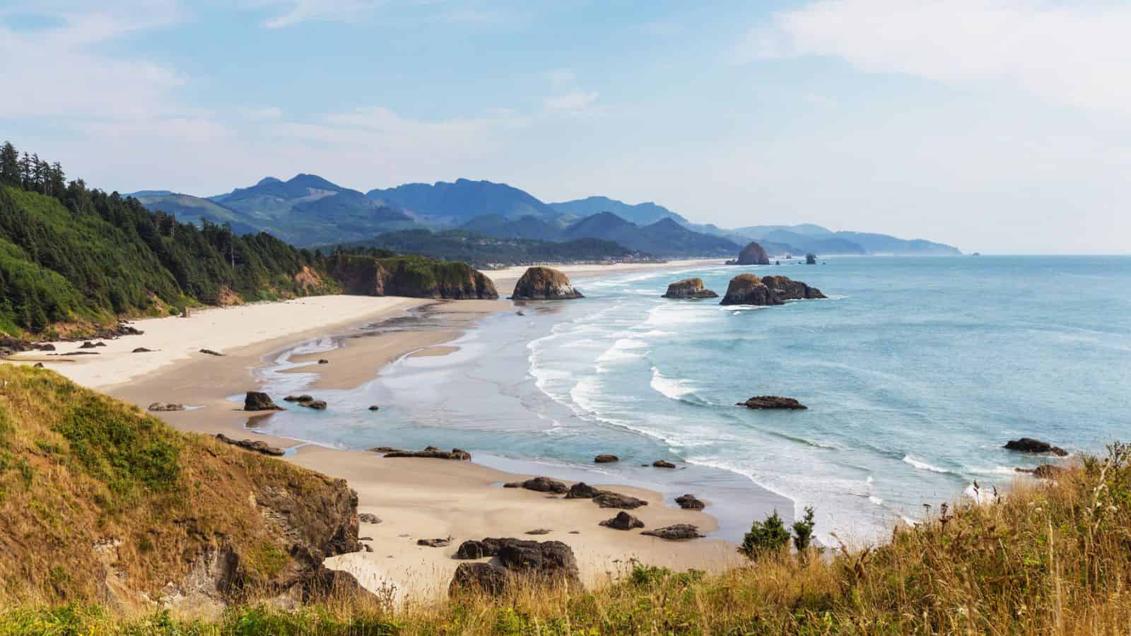 Sandy beach with rock formations, ocean waves, and forested hills under a partly cloudy sky.