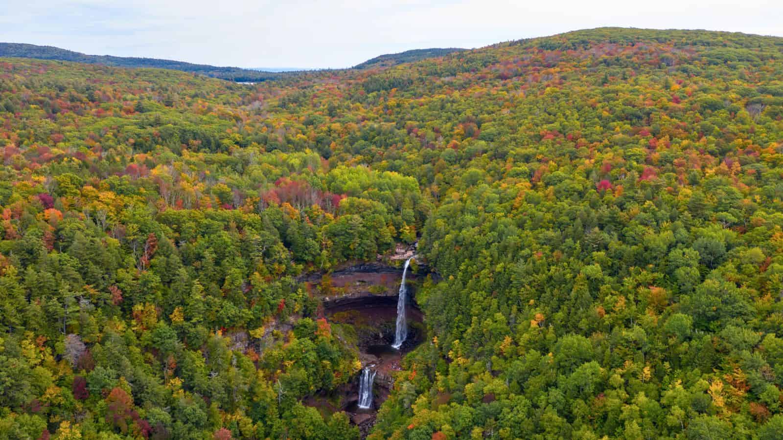 Aerial view of a tall waterfall surrounded by dense, colorful autumn forest on rolling hills.