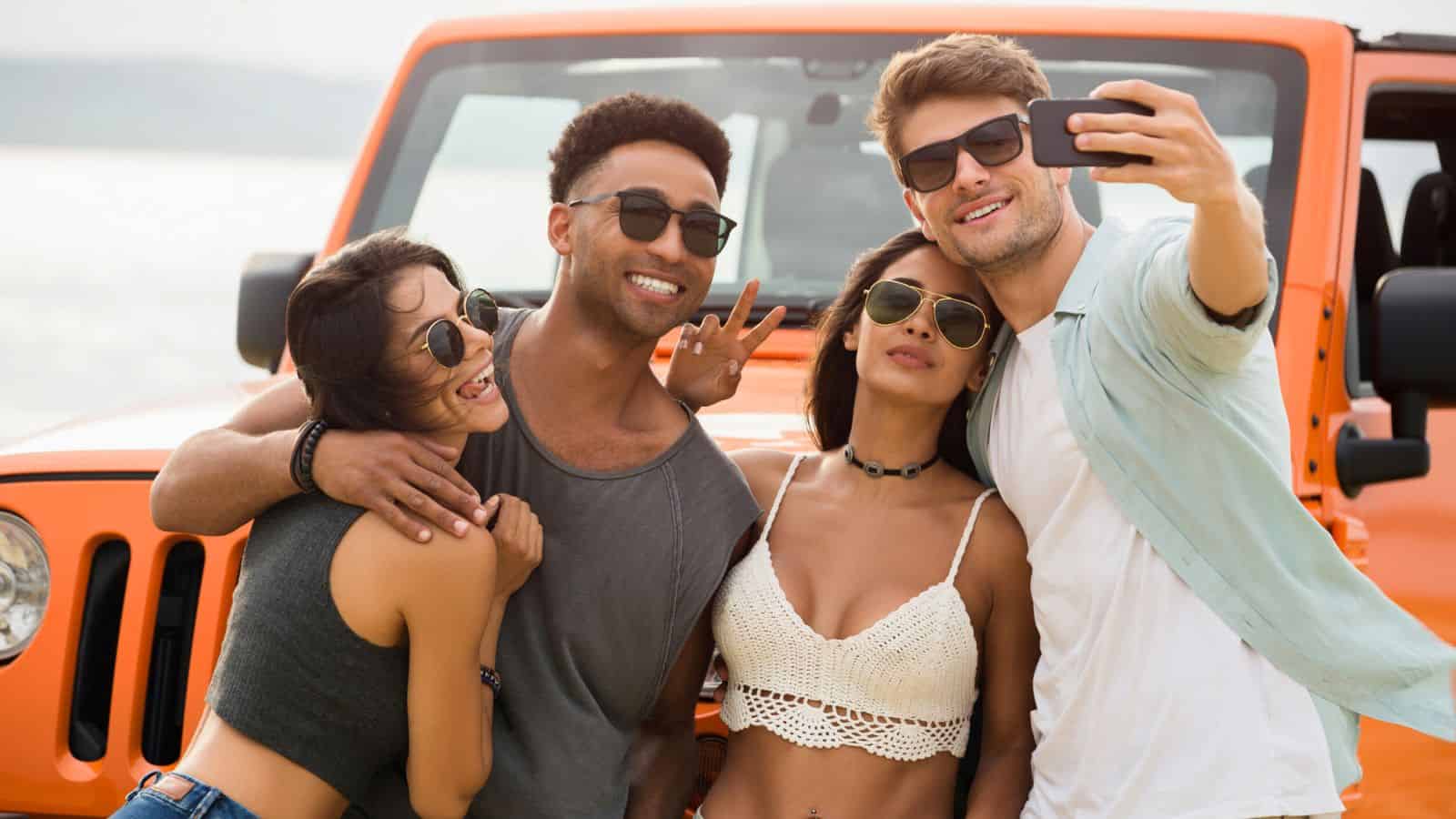 Four friends in sunglasses take a selfie in front of an orange jeep on a sunny day.