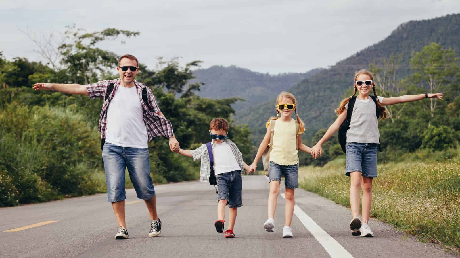 A man and three children in sunglasses walk hand in hand on a road with mountains in the background.
