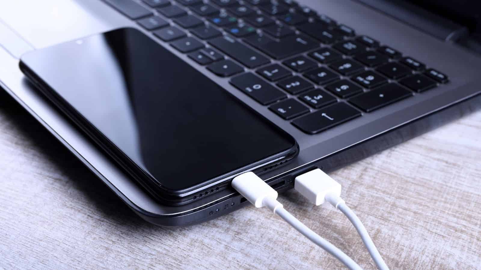 A smartphone charging with a USB cable connected to a laptop on a wooden surface.