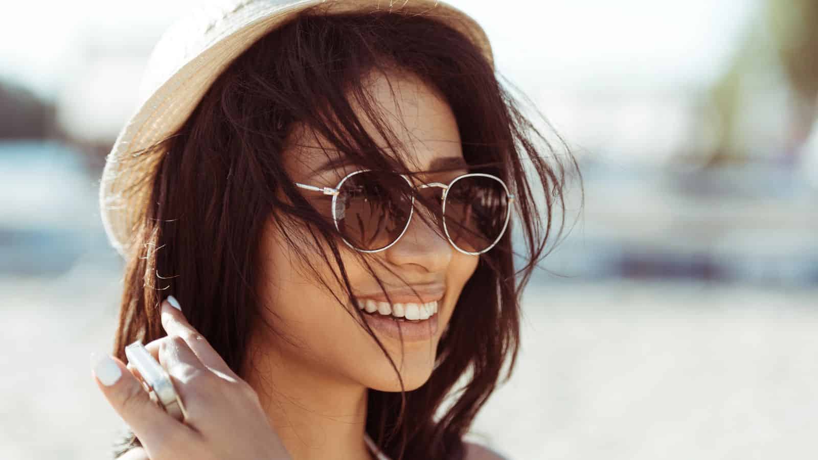 Smiling woman wearing sunglasses and a hat with windblown hair at the beach.