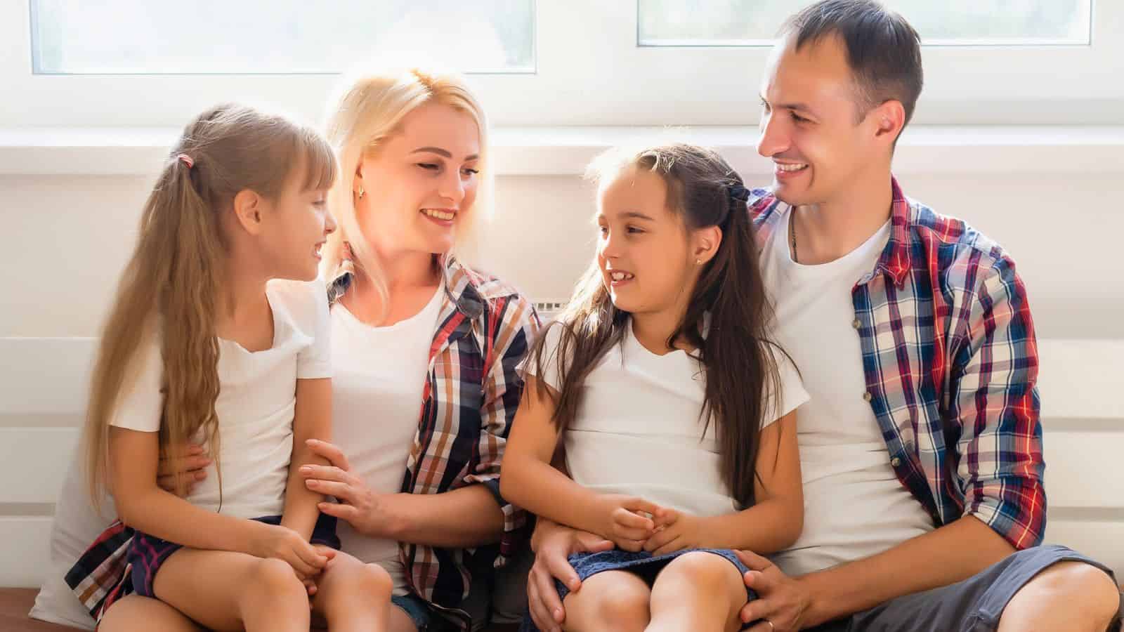 A smiling family of four sits together on a bench, wearing casual clothes and looking at each other.