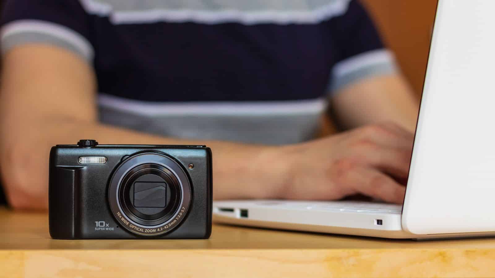 A digital camera on a table in front of a person using a laptop.