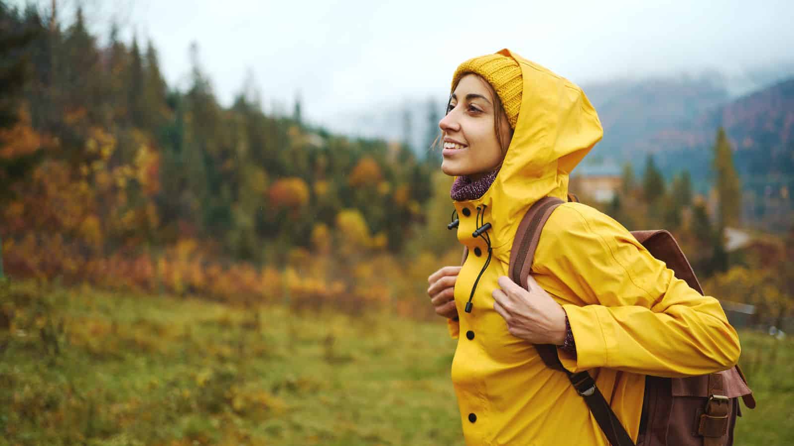 Person in a yellow raincoat and backpack smiling outdoors in a green, forested area with autumn colors.