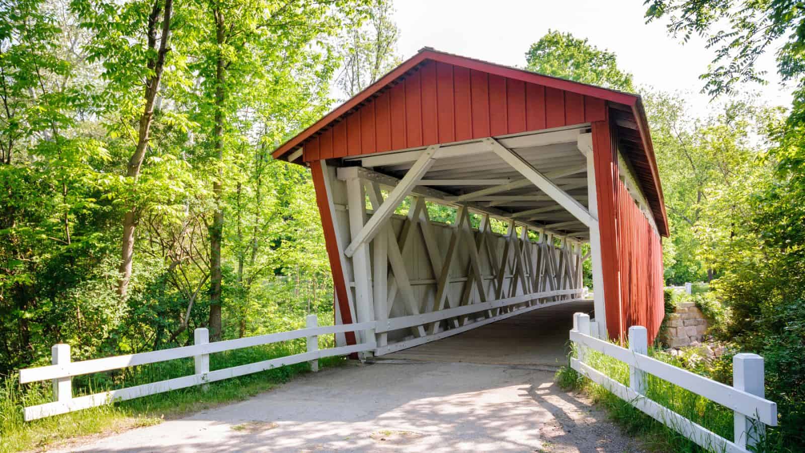 Red covered wooden bridge with white railings surrounded by green trees on a sunny day.