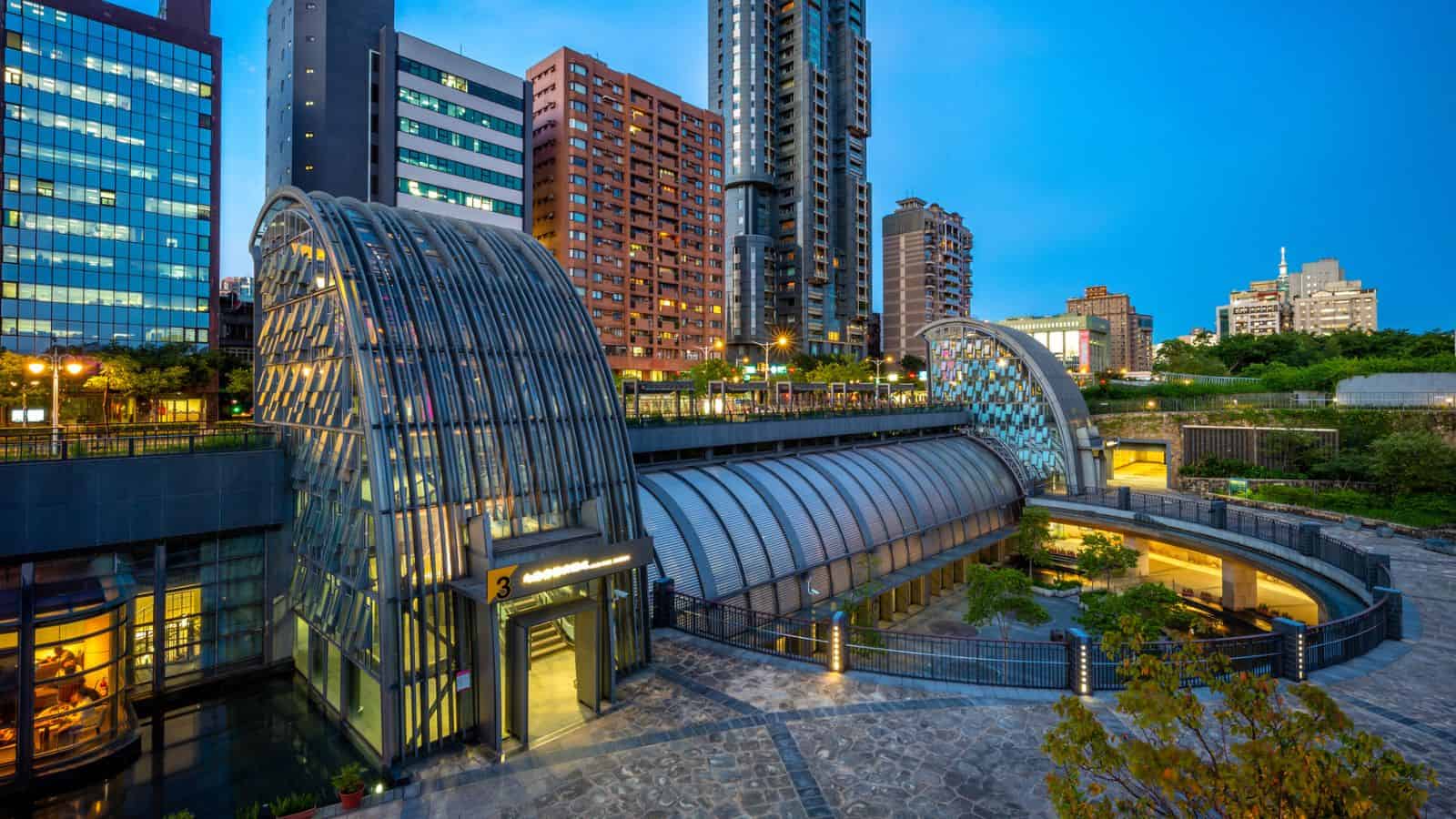 Modern urban buildings and a curved glass metro station entrance at dusk with city lights illuminating the area.