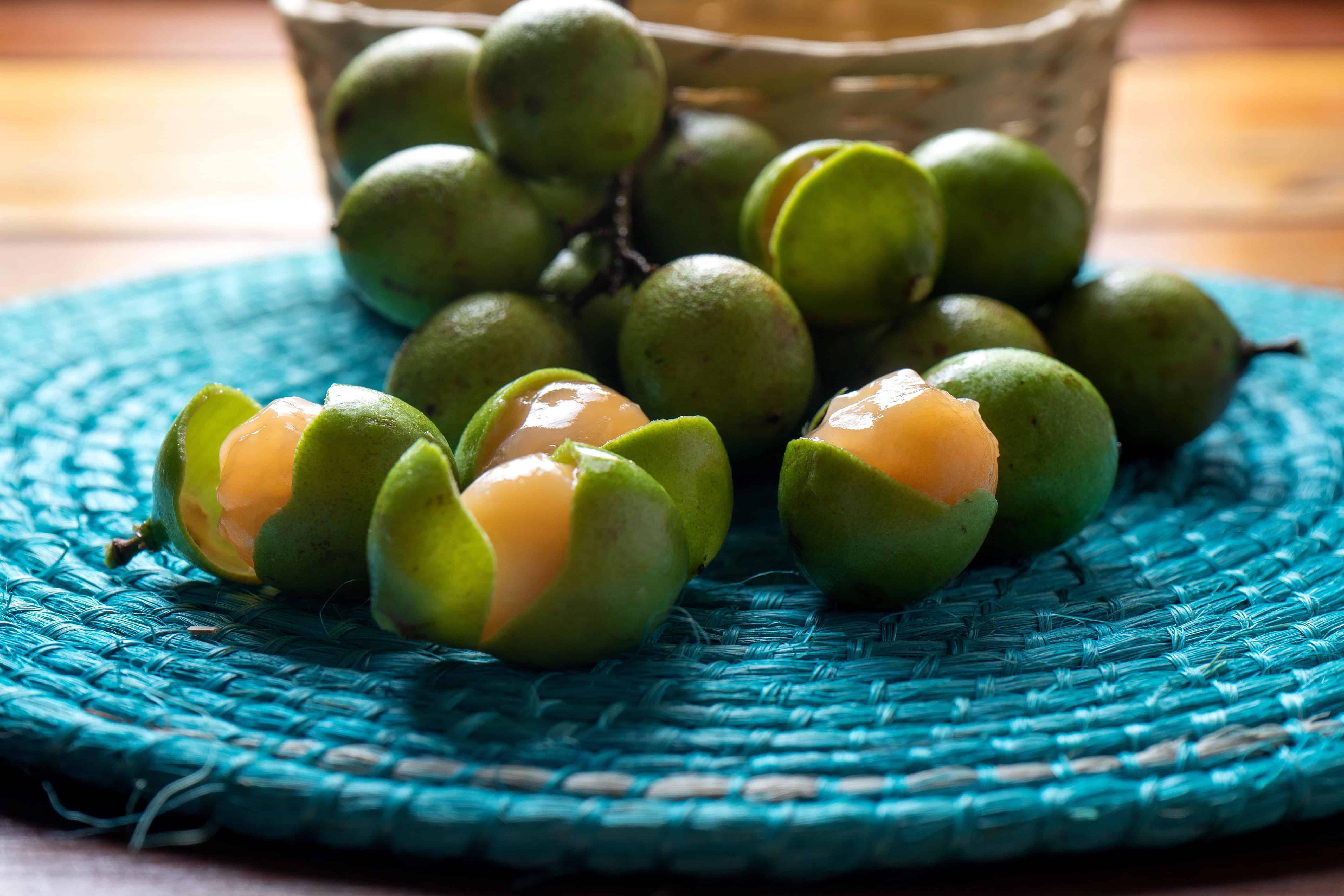 Green mamoncillo fruits, some peeled to reveal light orange flesh, rest on a blue woven mat with a basket in the background—an inviting display of exotic Colombian fruits.