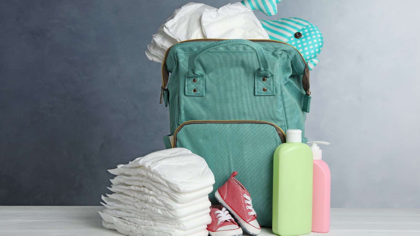 A diaper bag with travel essentials—diapers, baby lotion, a toy, and red baby shoes—sits on a table against a gray background, perfect for flying with kids or outings with kids under 5.
