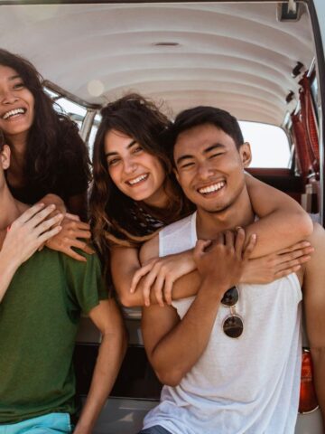 Four friends smile and pose together at the back of a van by the beach on a sunny day.