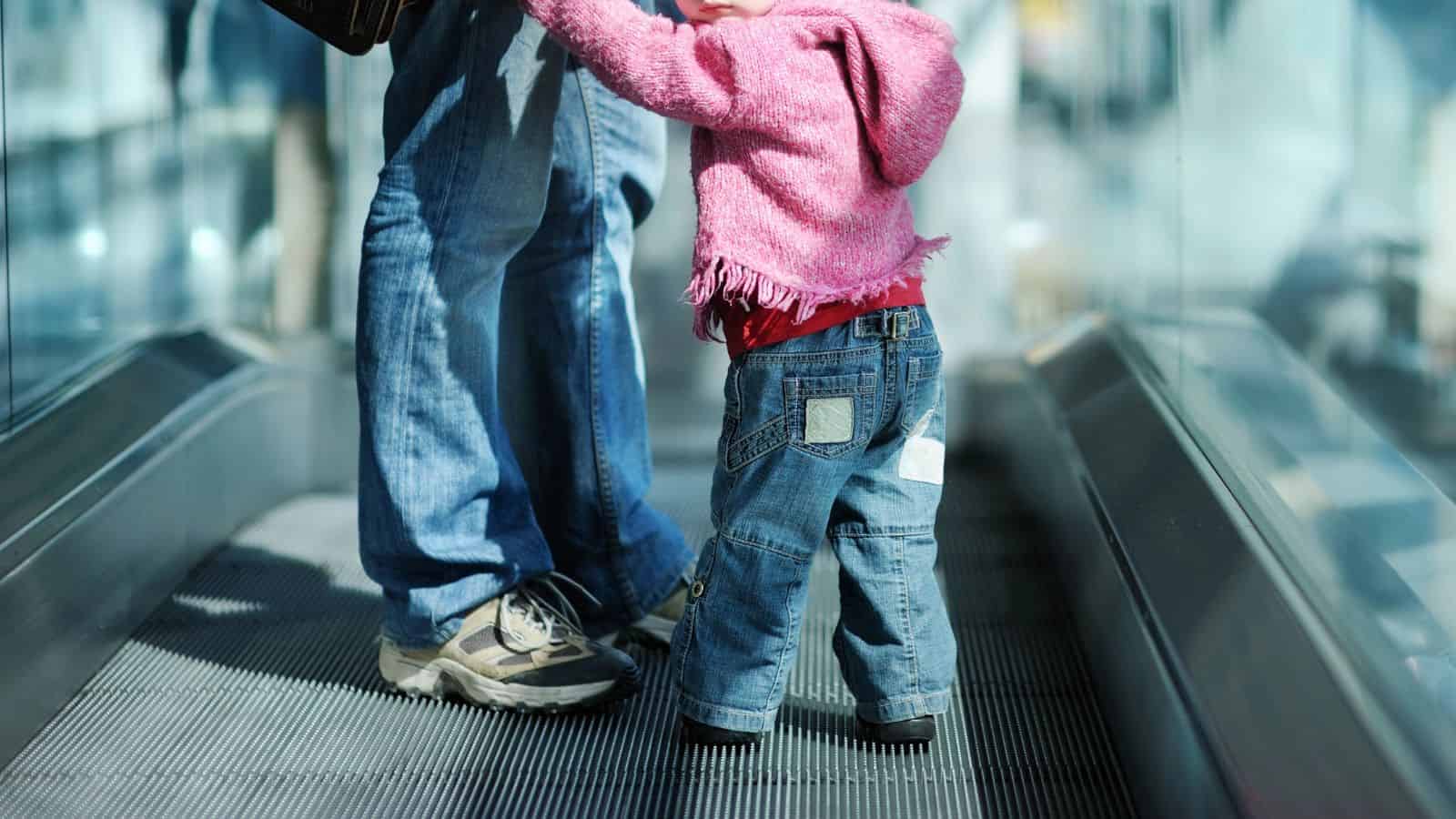 A toddler in jeans and a pink hoodie stands next to an adult on a moving walkway.