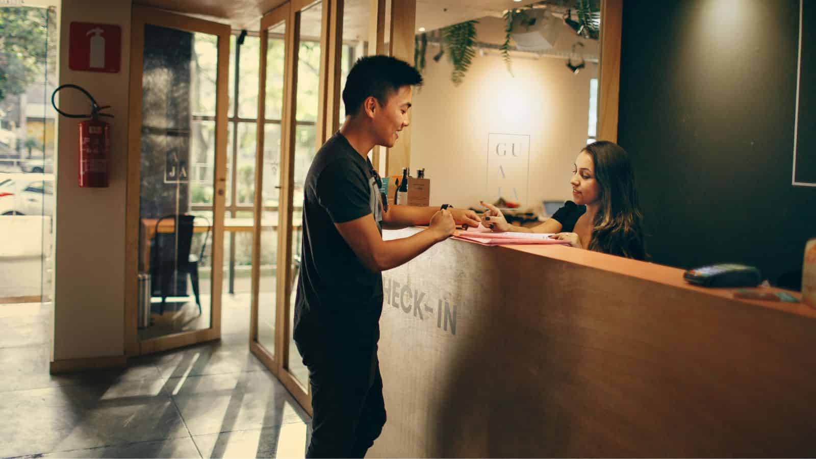 A man checks in at a reception desk while a woman behind the counter assists him with paperwork, carefully explaining any hidden fees related to his holiday travel.