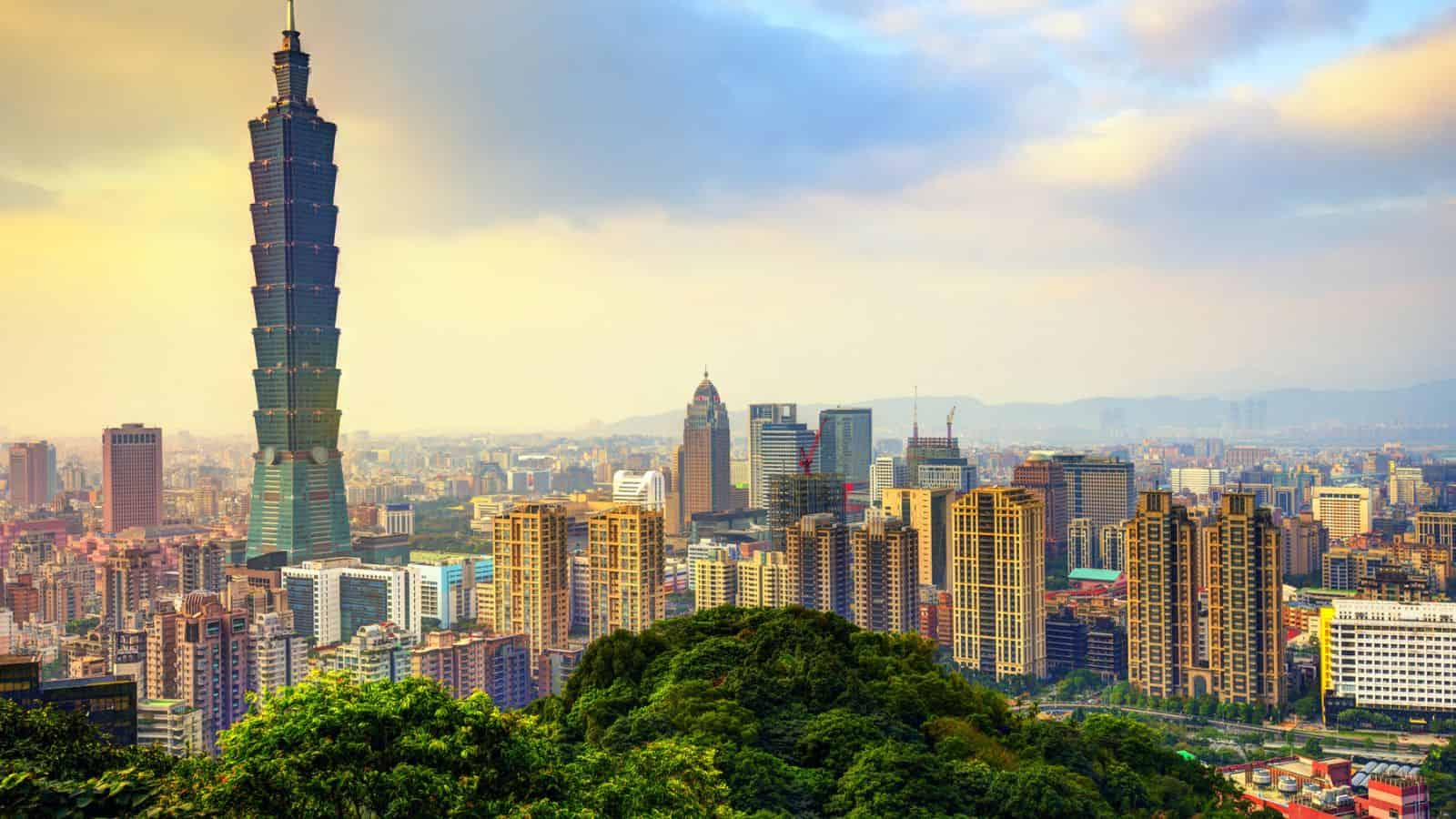 Taipei city skyline with Taipei 101 skyscraper and green hills in the foreground under a cloudy sky.