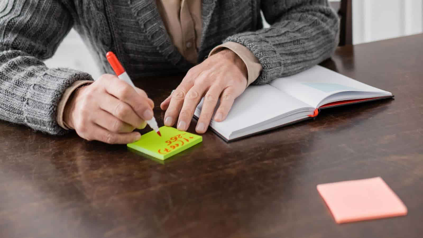 Person writing on a green sticky note with a red marker next to an open notebook on a wooden table.
