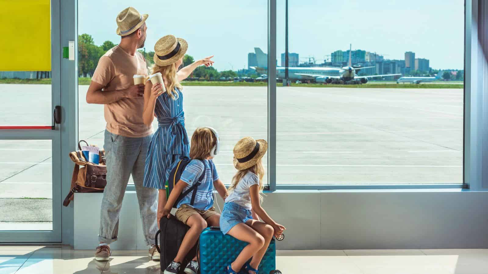 Family with hats and suitcases watching airplanes through airport window; woman points toward the runway.