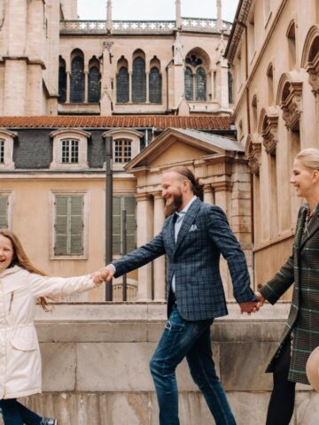 A family of three walks happily in front of a historic building with large windows and a rose window.