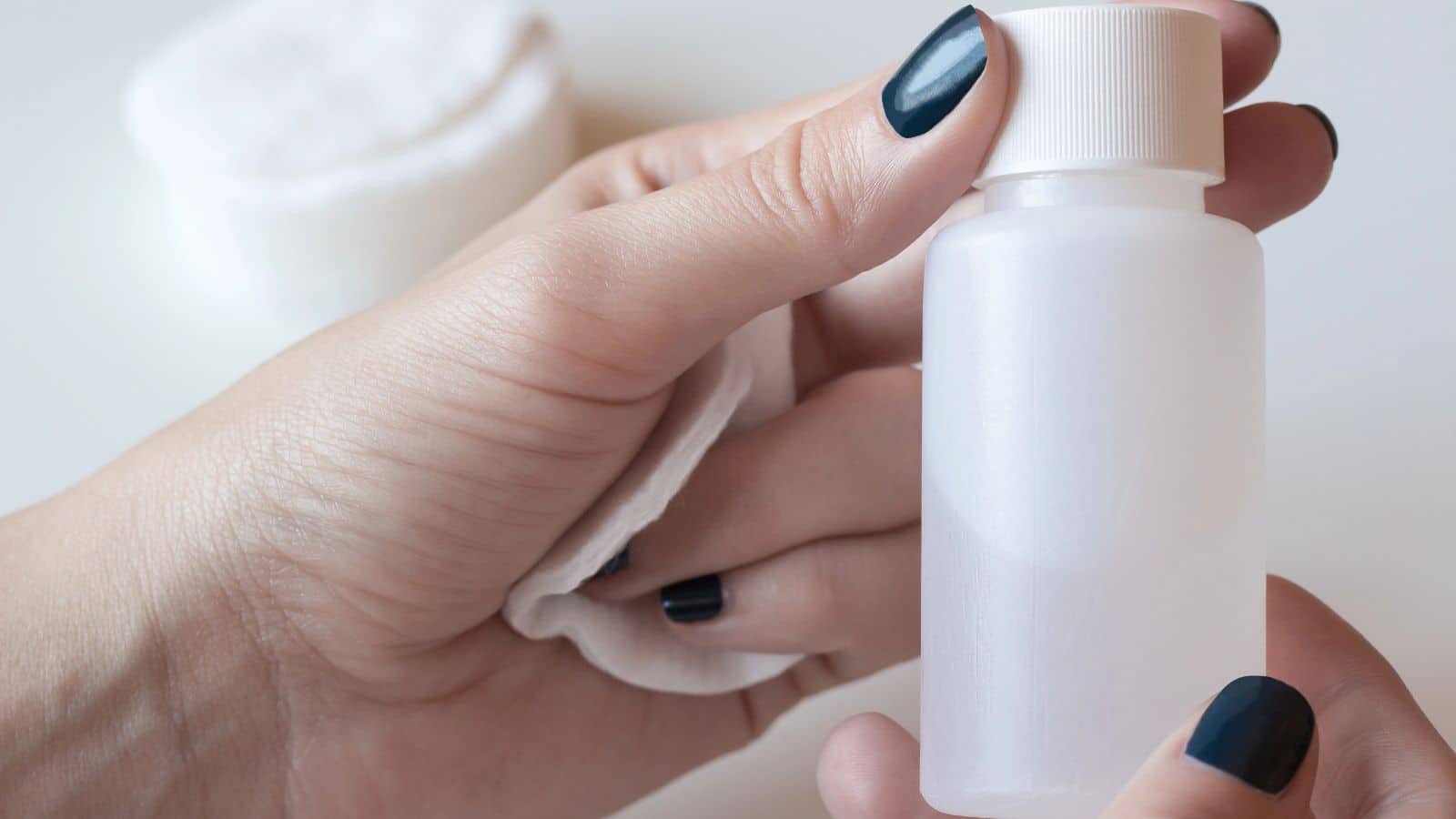 A hand holding a white bottle and cotton pad, with dark-painted nails visible.