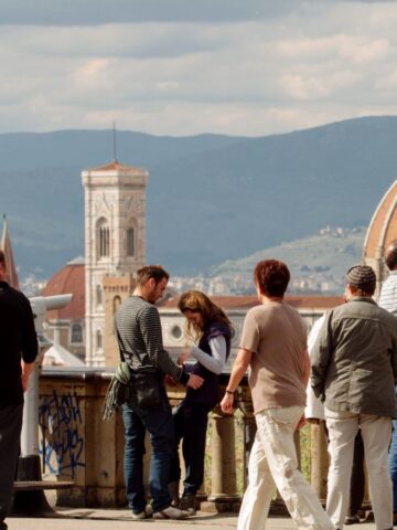 Tourists take photos of Florence’s Duomo and bell tower from a scenic overlook on a sunny day.