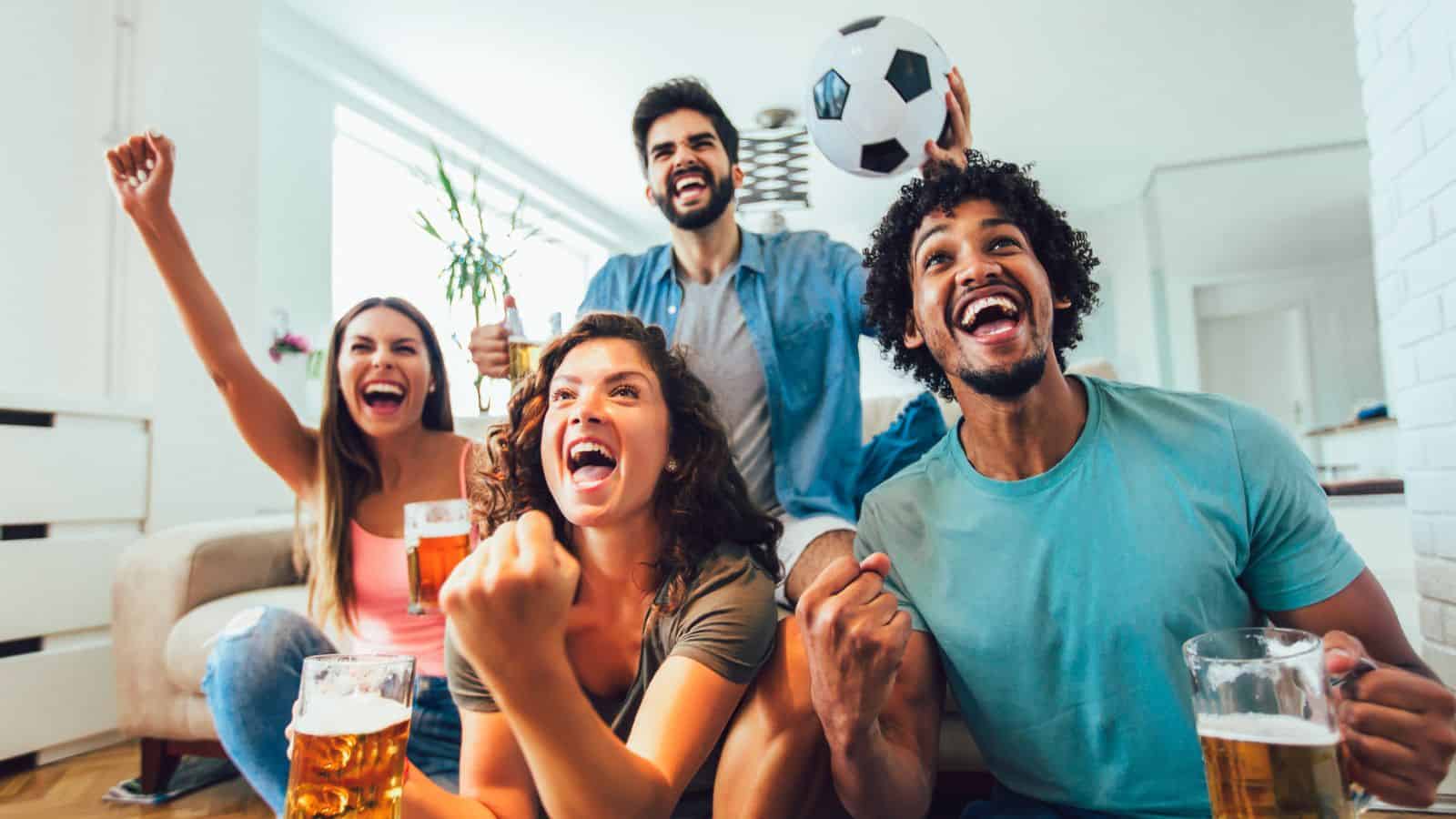 Four friends cheer and celebrate while watching a soccer game at home, holding drinks and a soccer ball.