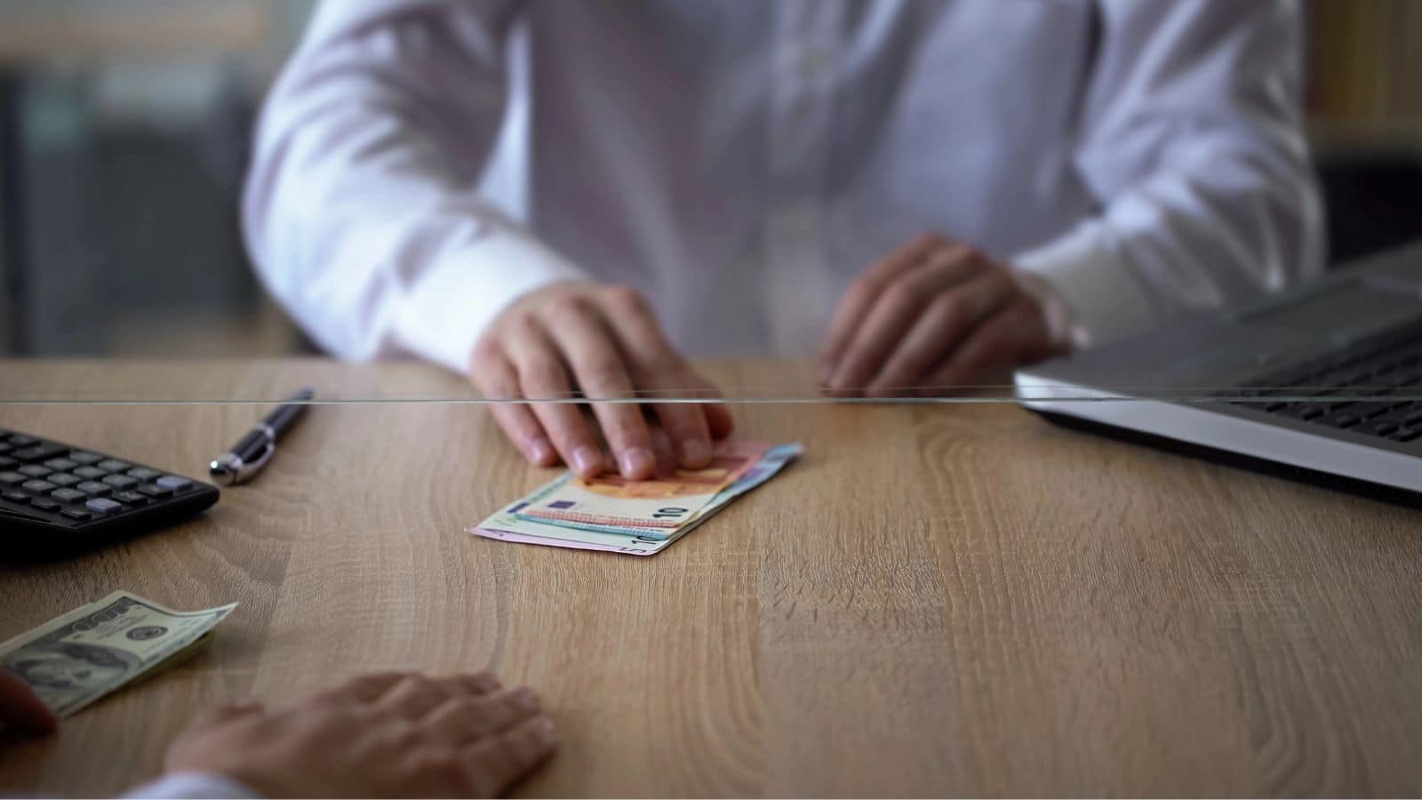 Person in white shirt counting euro bills at a desk with a laptop, pen, calculator, and some dollars.