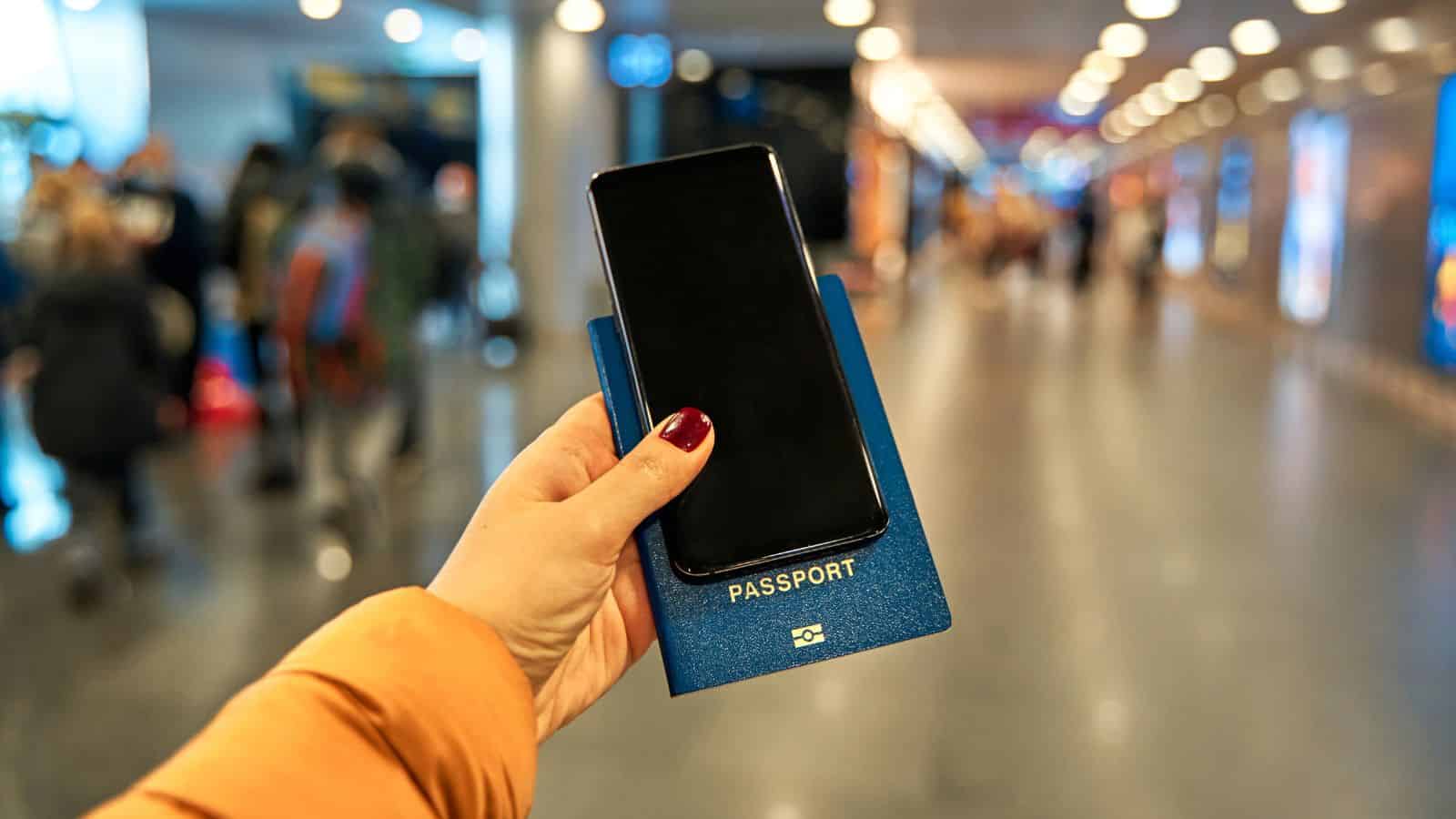 Person holding a smartphone and passport in an airport terminal with people in the background.