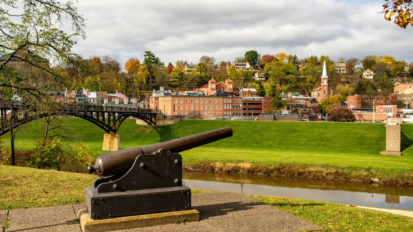 Historic cannon overlooks a river with a bridge and small town buildings on a hill with autumn trees.