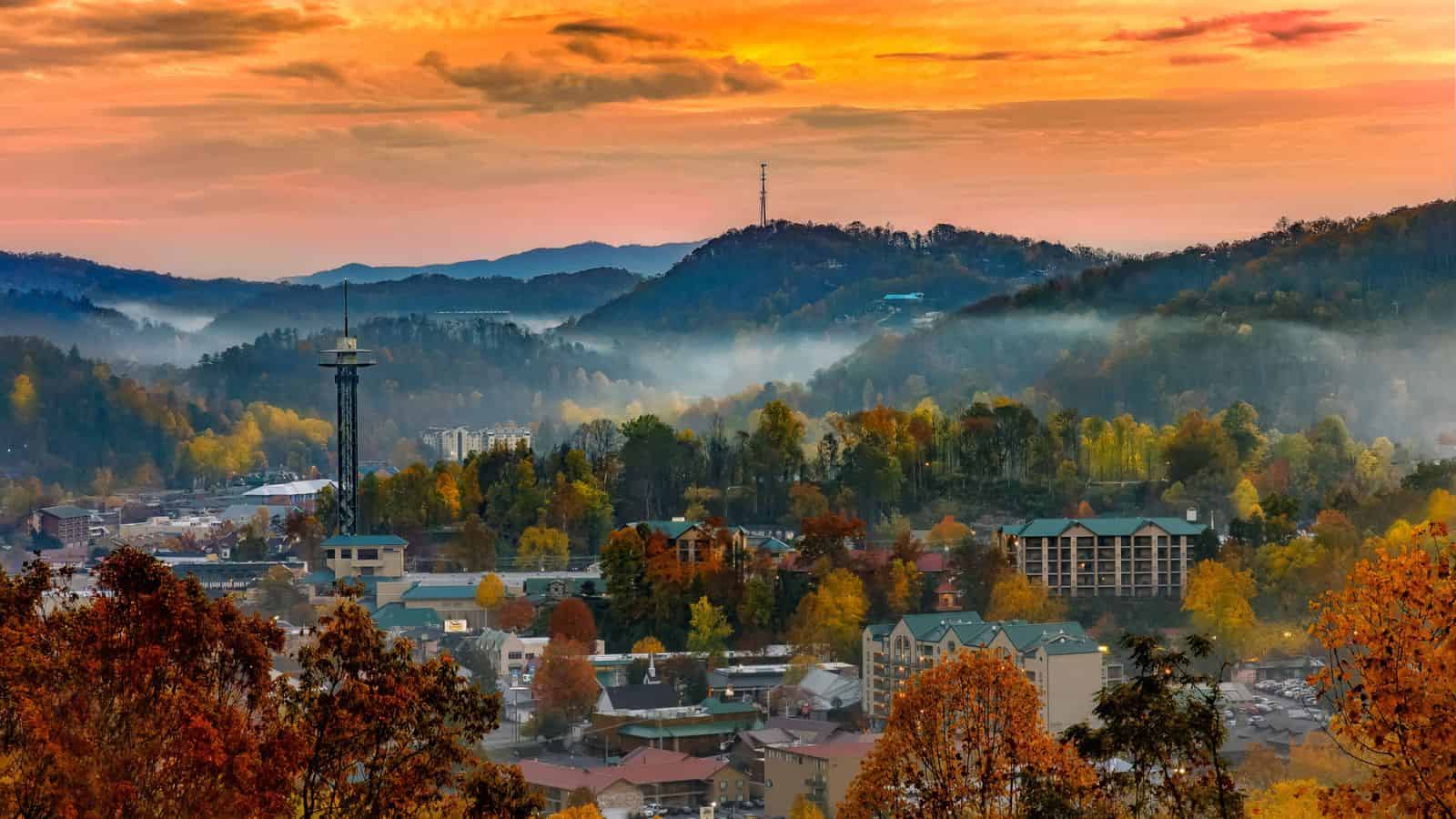 Aerial view of a town in a valley with autumn trees, mist, and a bright orange sunset sky.