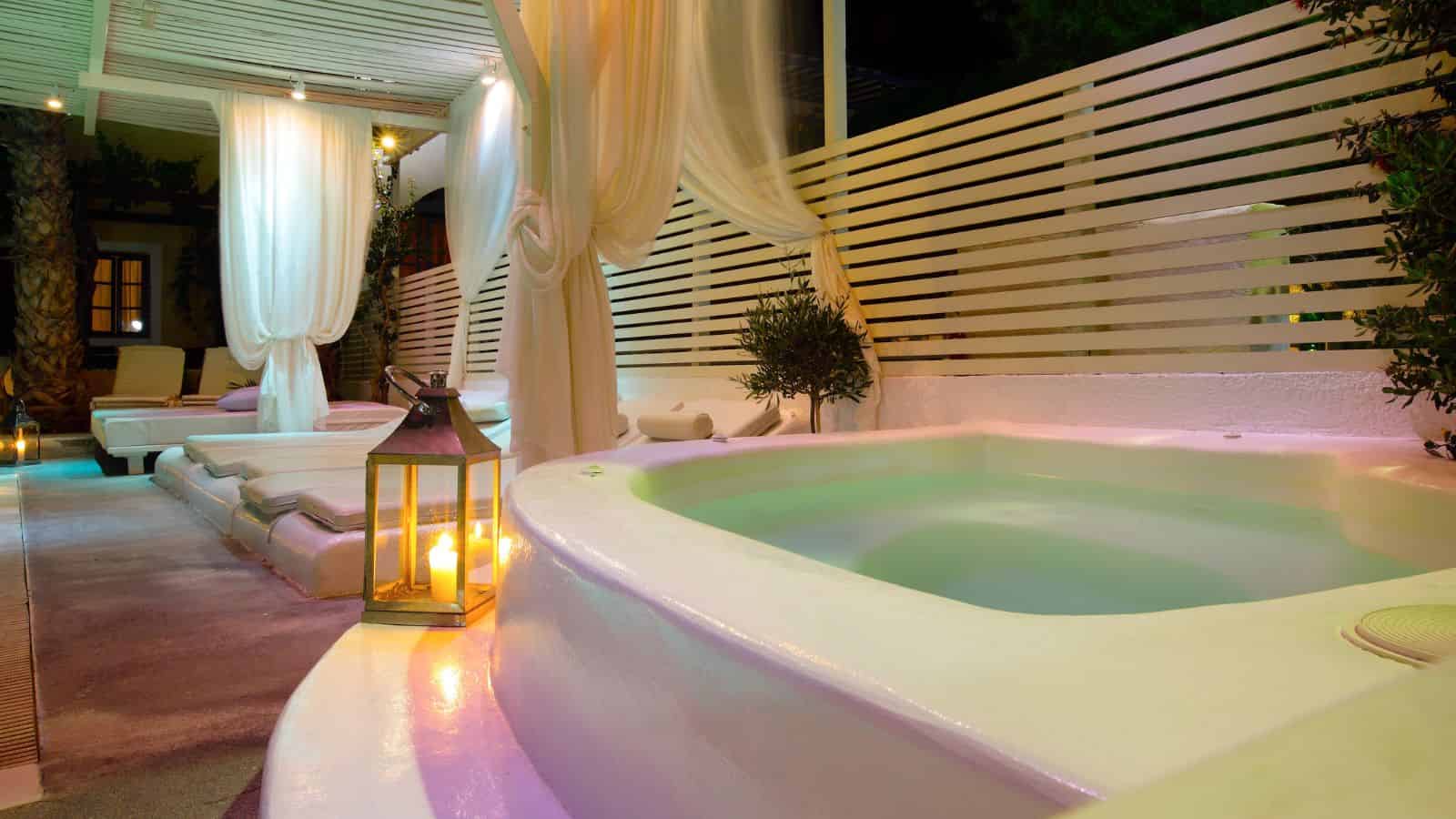 Outdoor spa with white lounge beds, draped curtains, a hot tub, and soft lantern lighting at night.