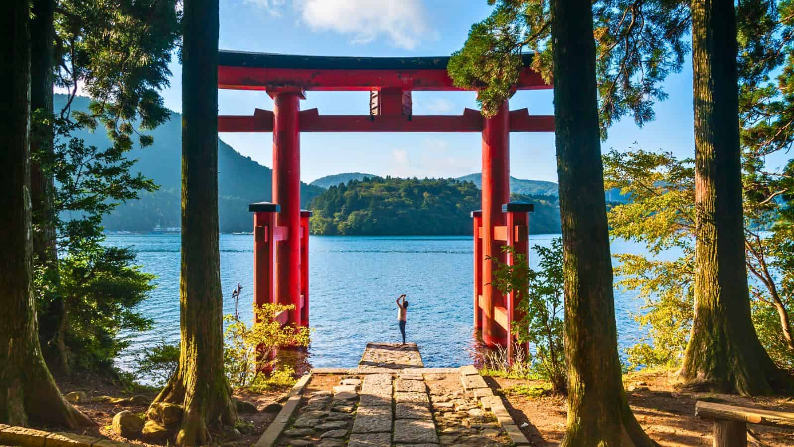 A person stands by a large red torii gate by a lakeshore, surrounded by trees and mountains.