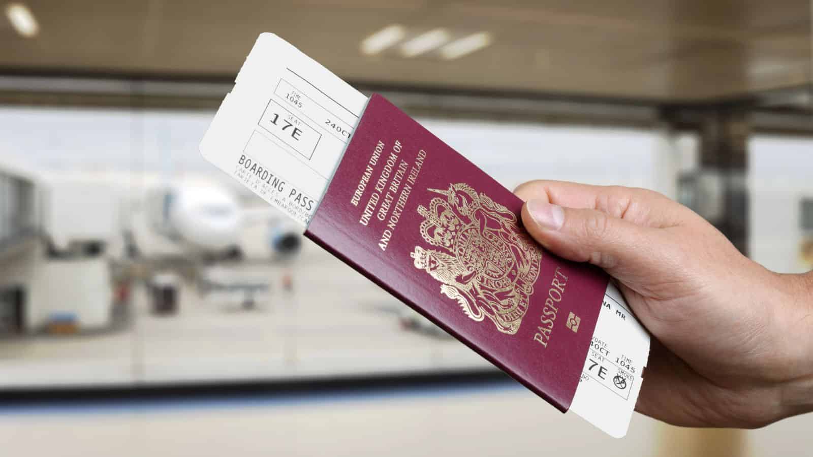Hand holding a UK passport and a boarding pass at an airport, with an airplane visible outside the window.