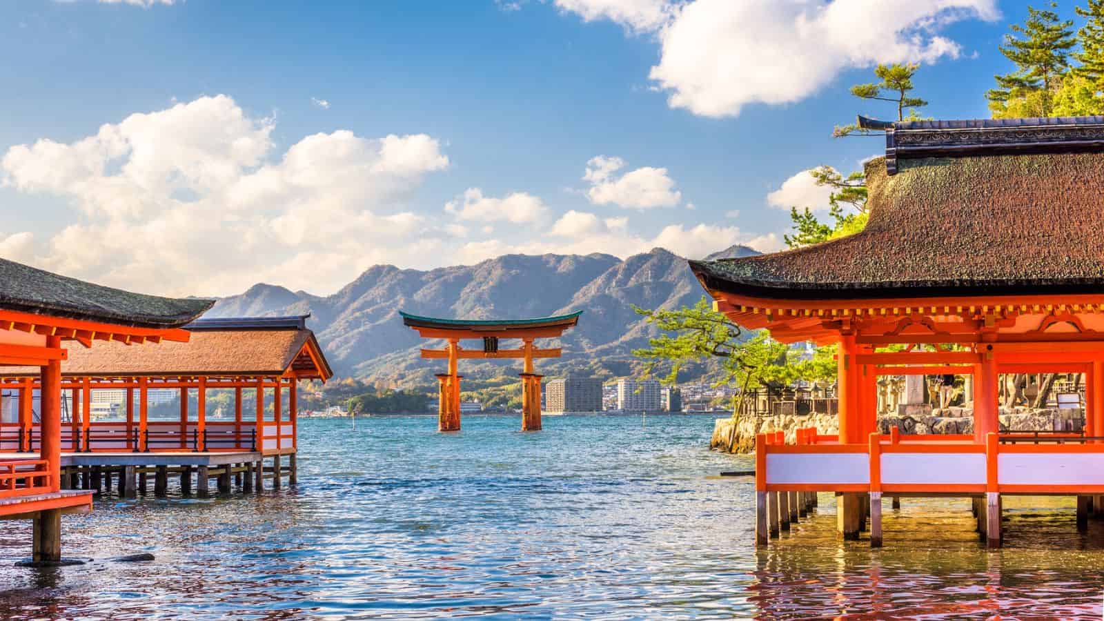 Red shrine buildings frame a torii gate in the water, with mountains and blue sky in the background.