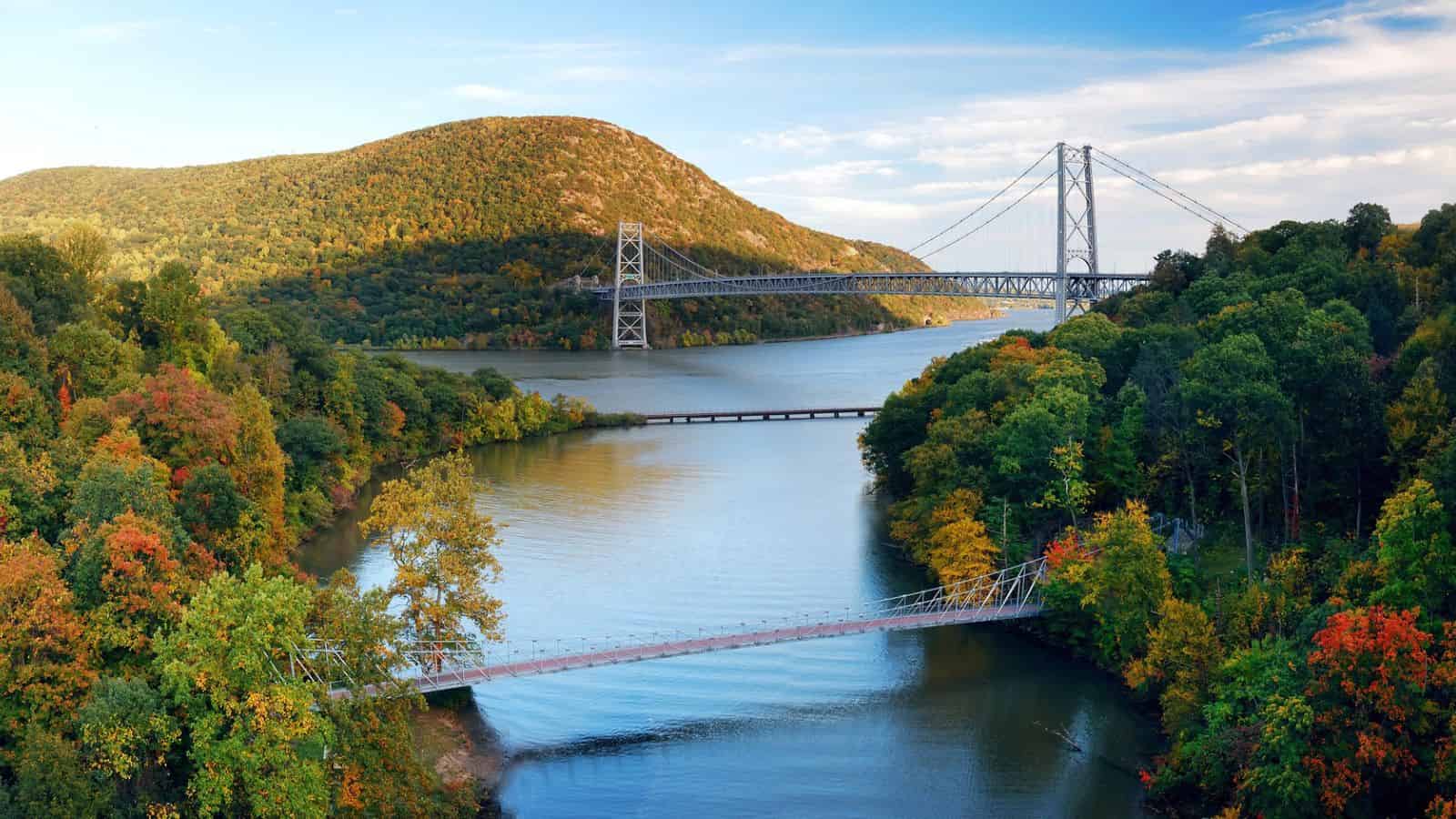 Three bridges span a river surrounded by green, yellow, and orange autumn trees and hills under a blue sky.
