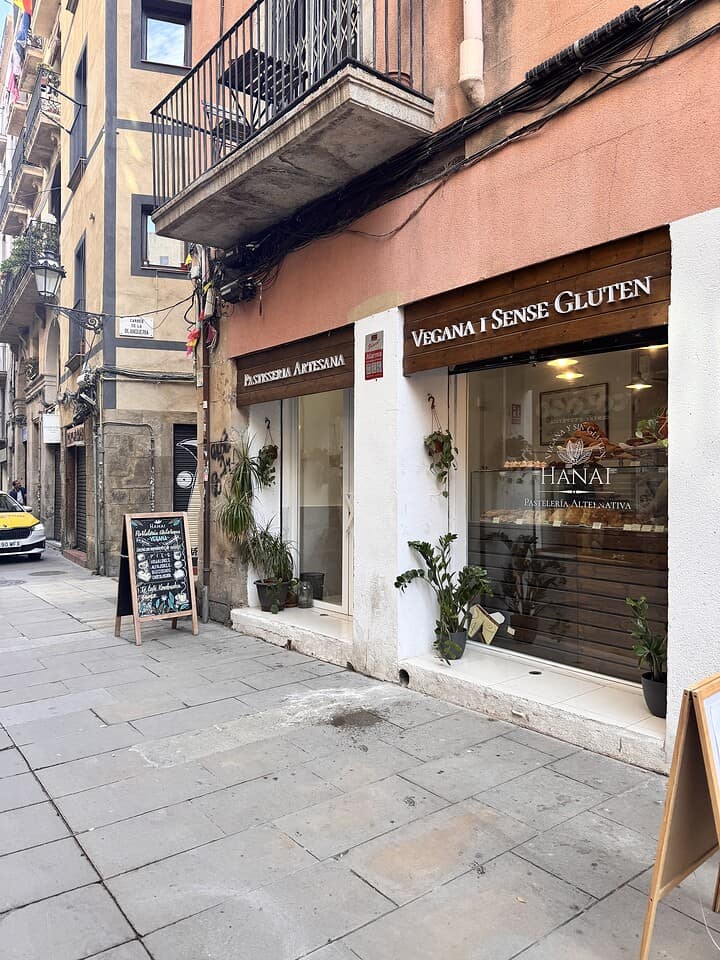 Bakery storefront with signs for vegan and gluten-free products, plants, and a menu board on a city street.