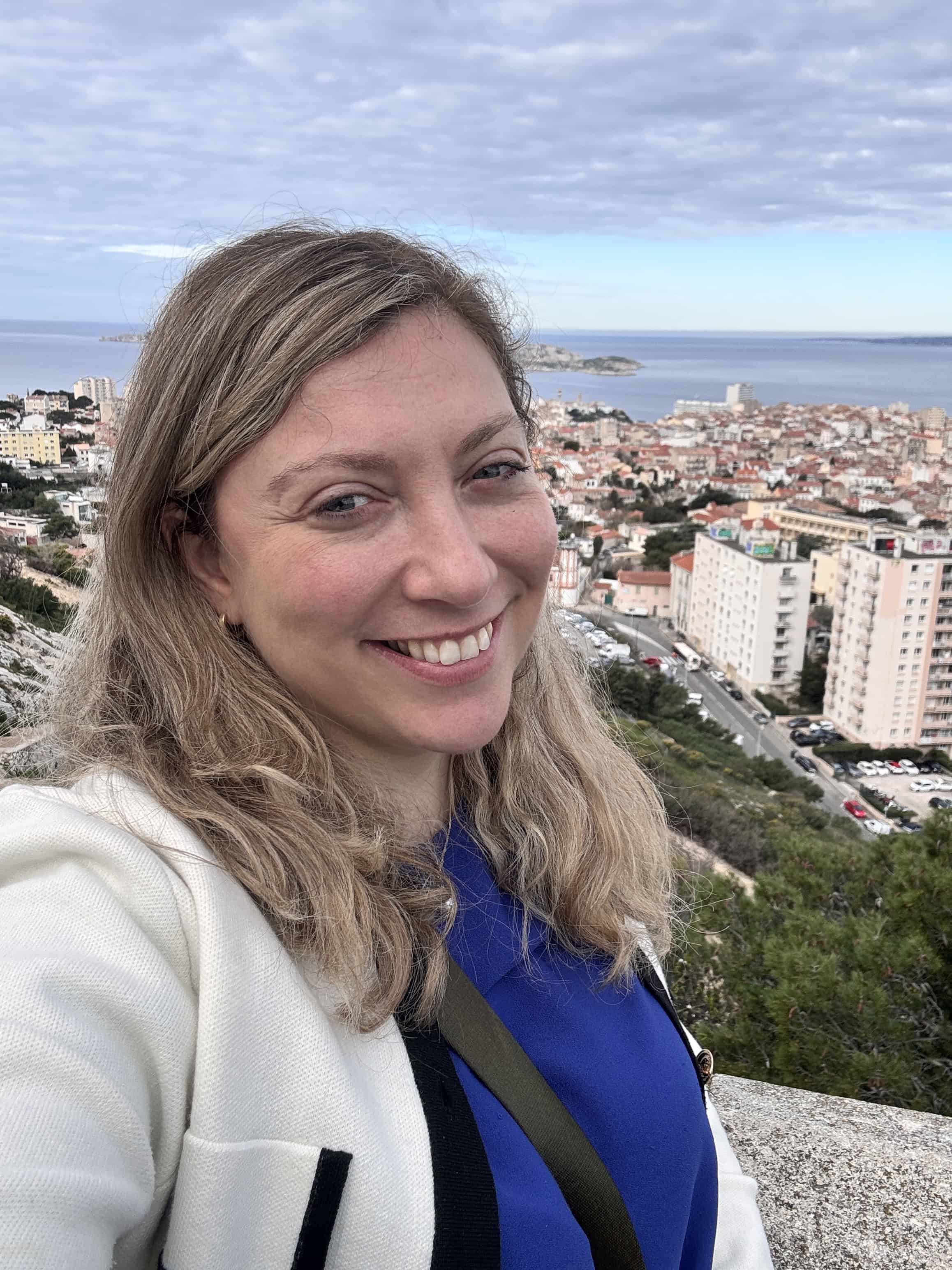 Woman smiling in selfie with a coastal city, ocean, and buildings in the background under a cloudy sky, capturing memories from her family’s 7-day itinerary aboard the MSC Fantasia Cruise.