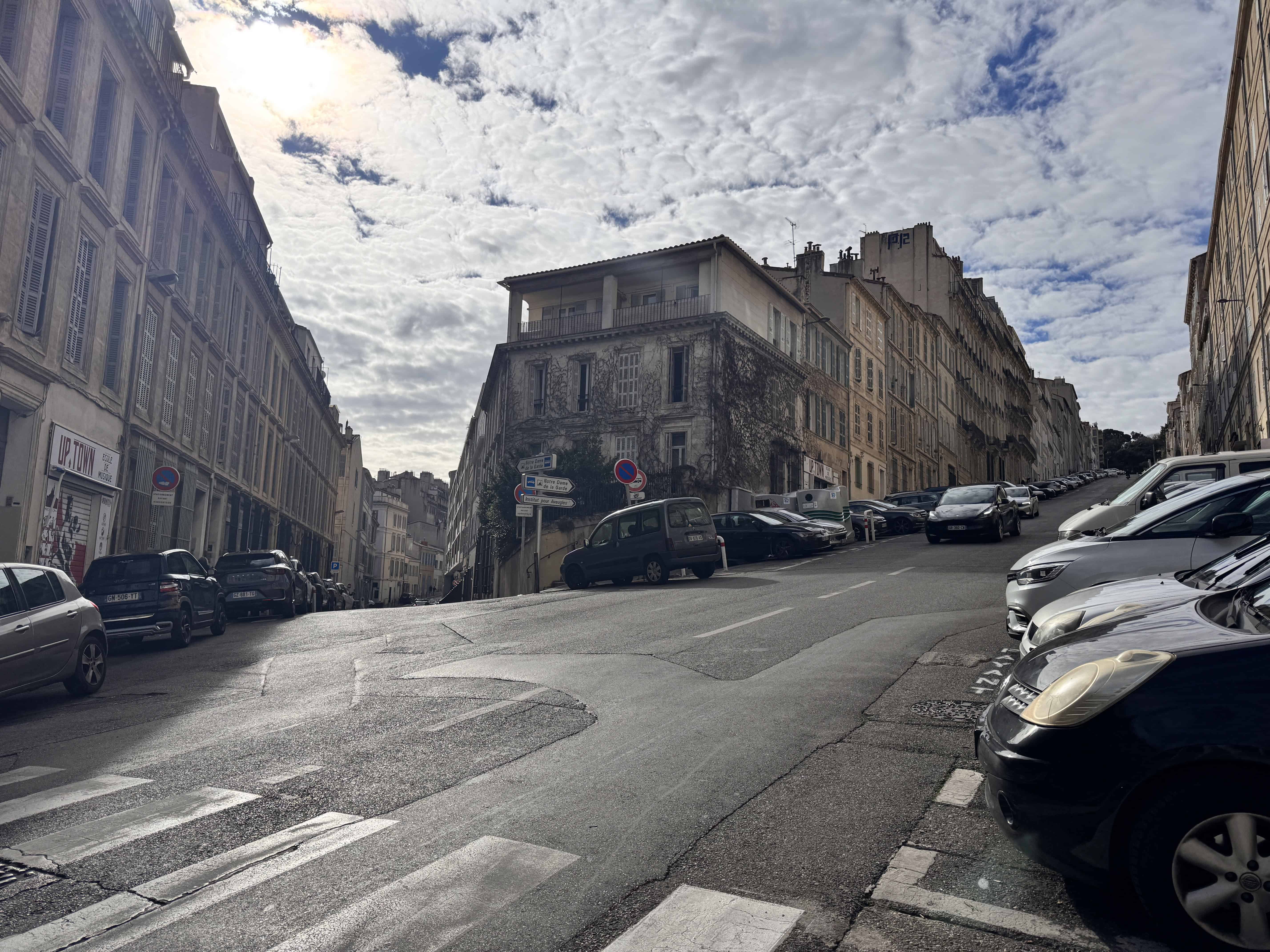 Steep city street with parked cars, old buildings, and a partly cloudy sky overhead.