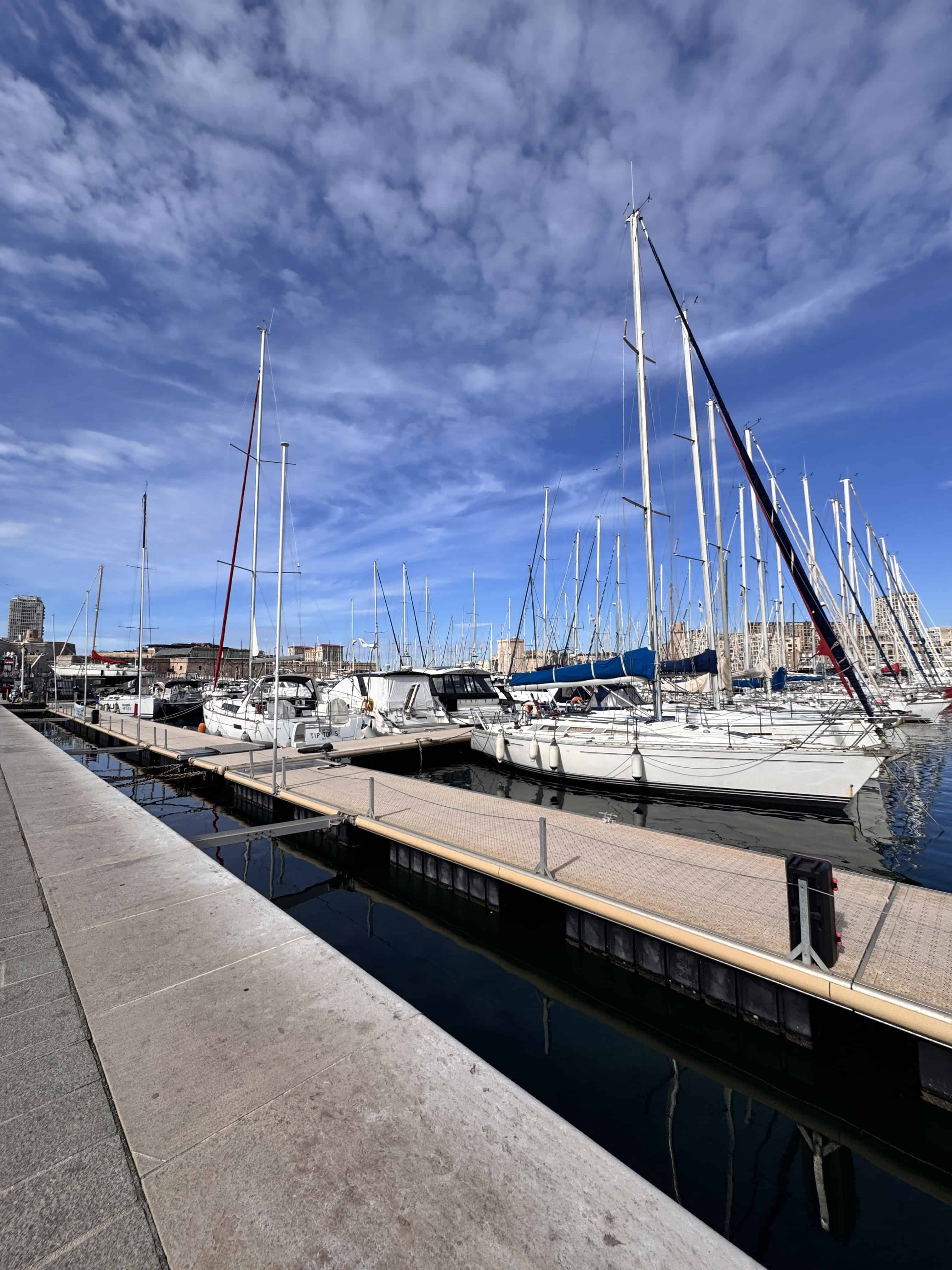 Sailboats are docked at a marina under a blue sky with scattered clouds.