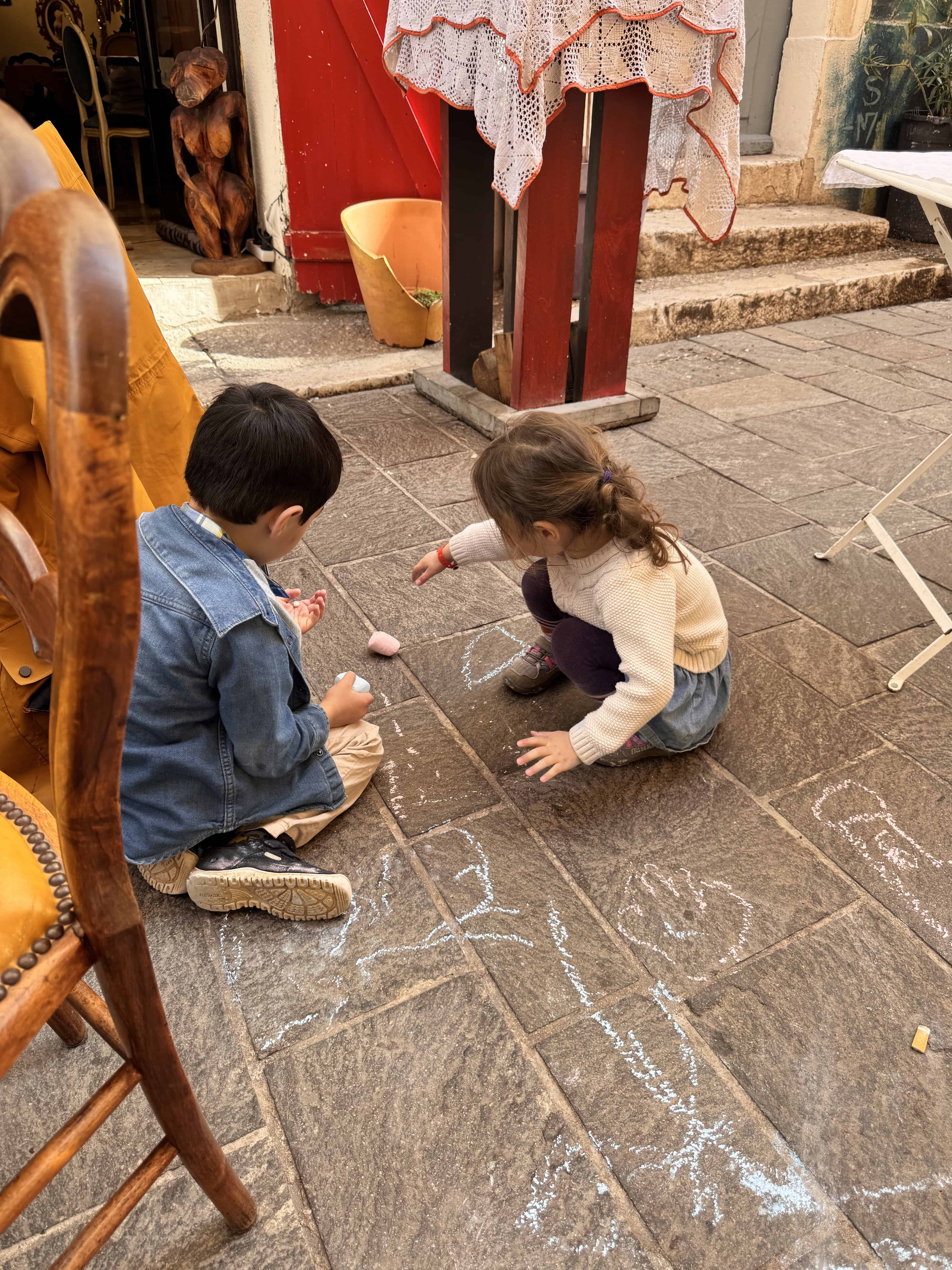 Two young children draw with chalk on stone pavement outside, surrounded by chairs and colorful objects, exchanging stories about their adventures and sharing first time cruise tips.