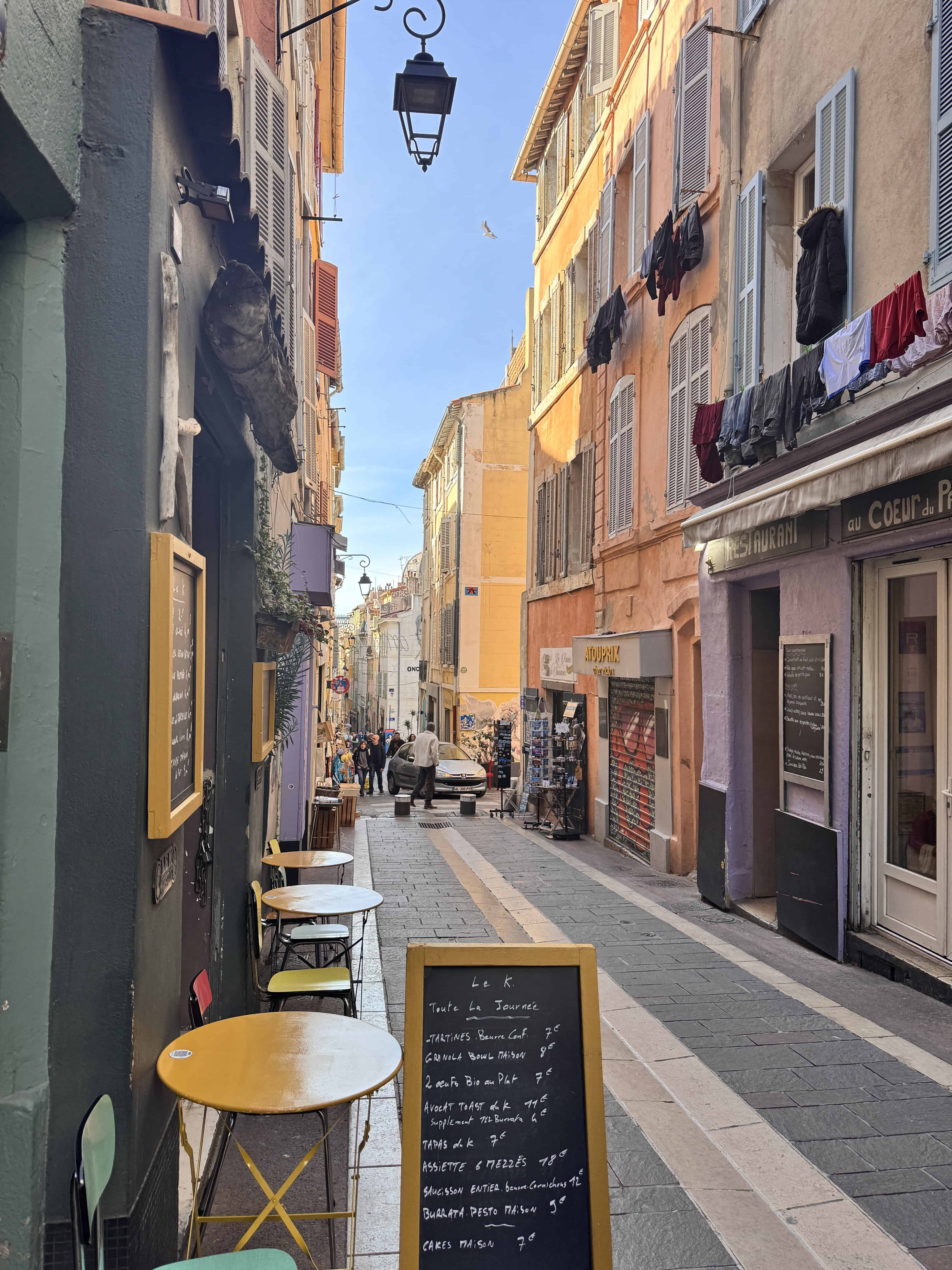 Narrow European street in Marseille with colorful buildings, outdoor café tables, and laundry hanging above—a perfect stop on your France itinerary.