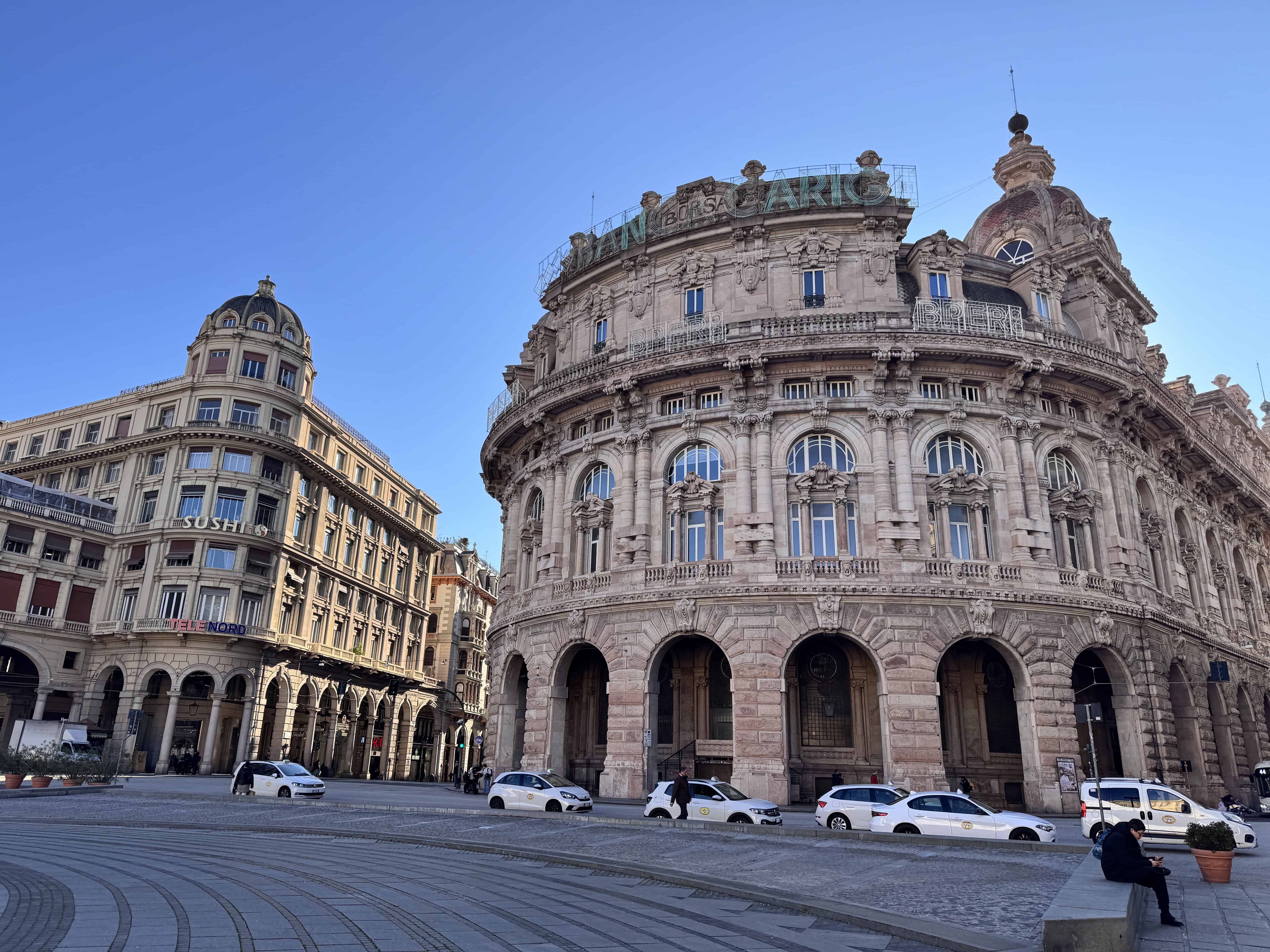 Historic stone buildings and parked white taxis in a sunny European city square with clear blue sky.