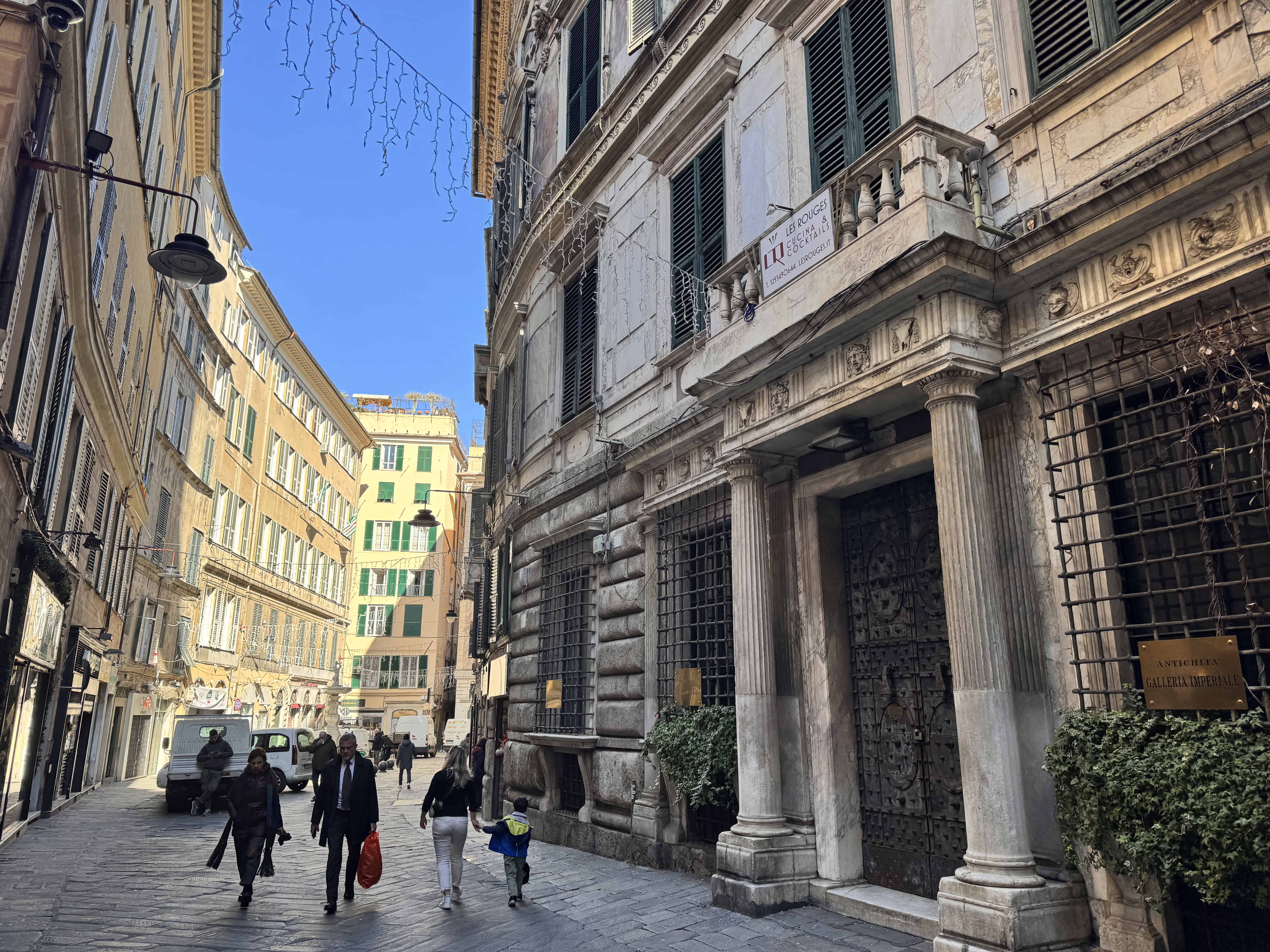 People walk down a narrow, sunlit street lined with old, ornate buildings on a clear day.