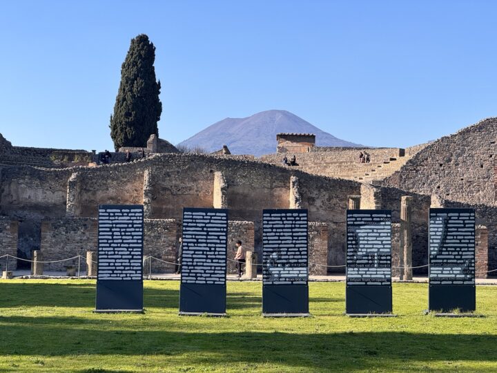 Five black panels with white markings stand before ancient ruins, with a mountain and blue sky in the background.