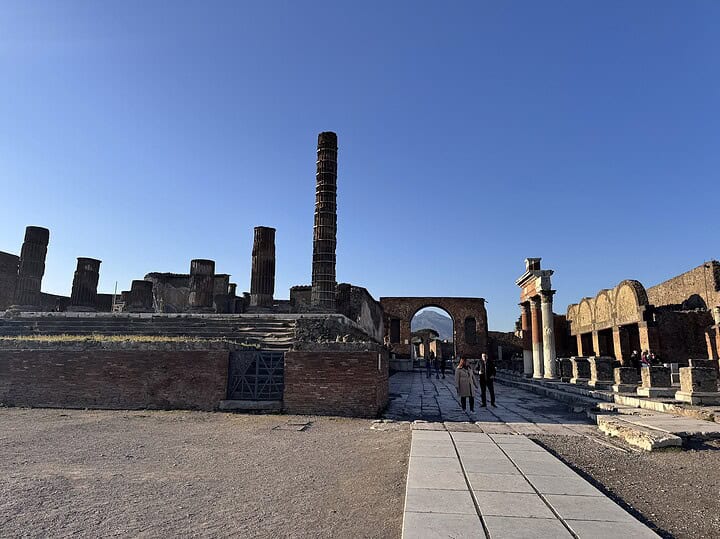 Ancient ruins under a clear blue sky with a tall column and a few people walking on a stone path.