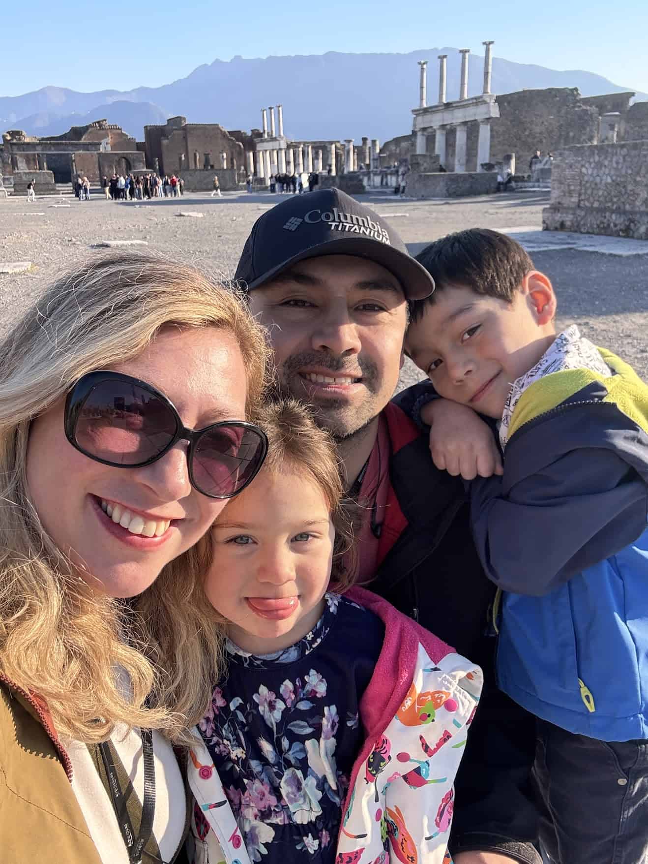 A smiling family of four poses for a selfie at ancient ruins on a sunny day.