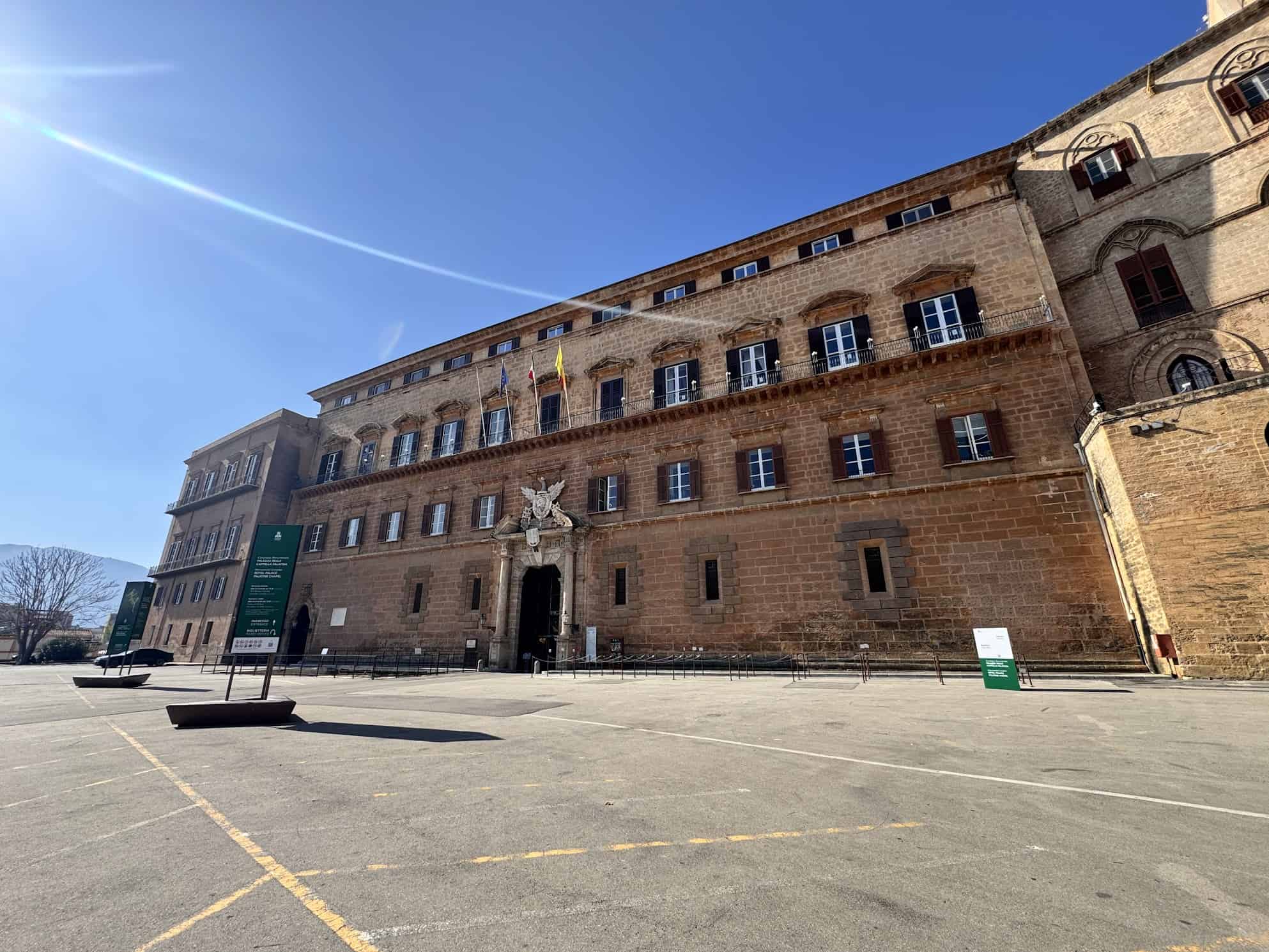 Large historic brown stone building with flags and a crest under a clear blue sky.